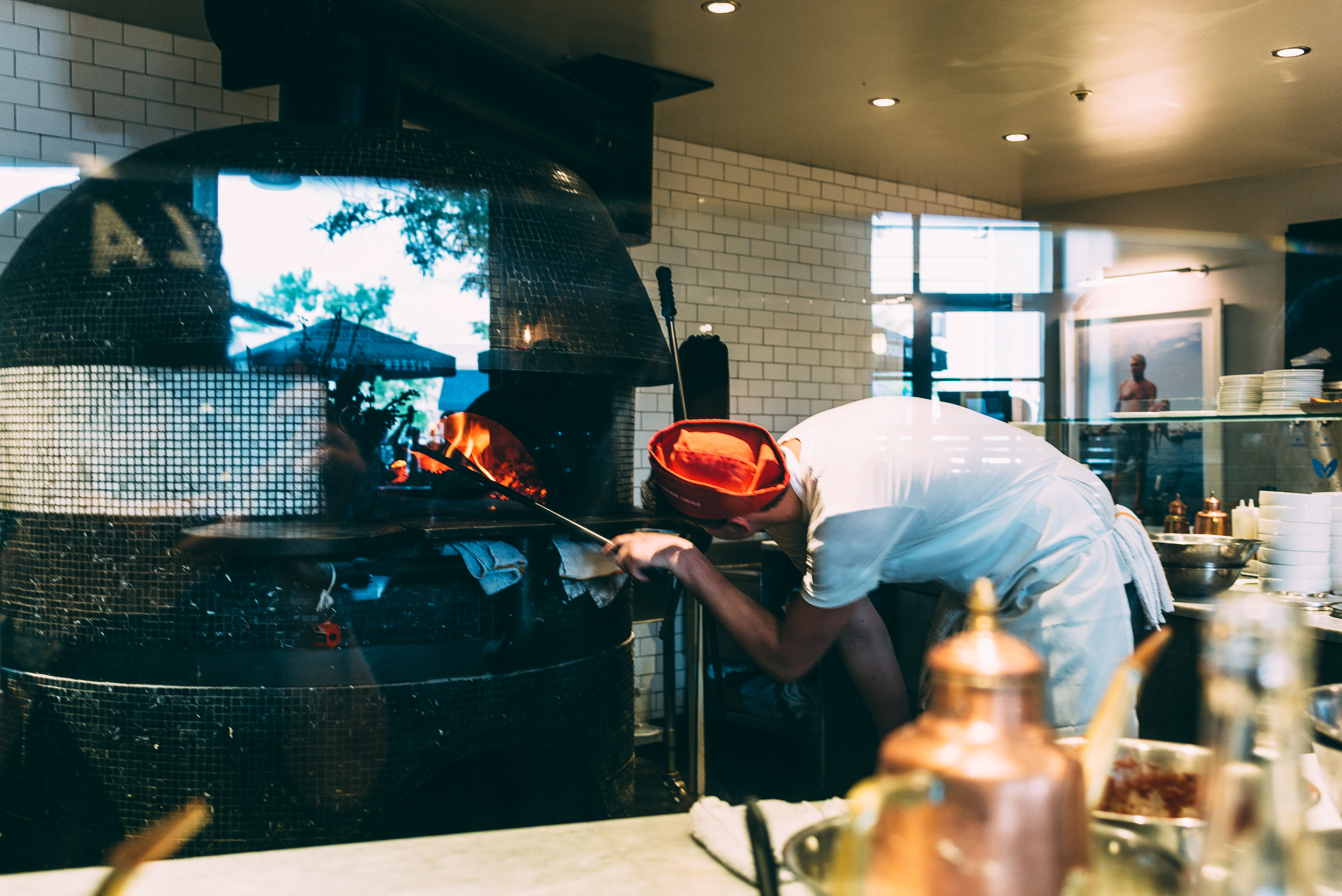 Pizza chef in a red cap tending a wood-fired pizza oven with glowing orange flames in a kitchen.