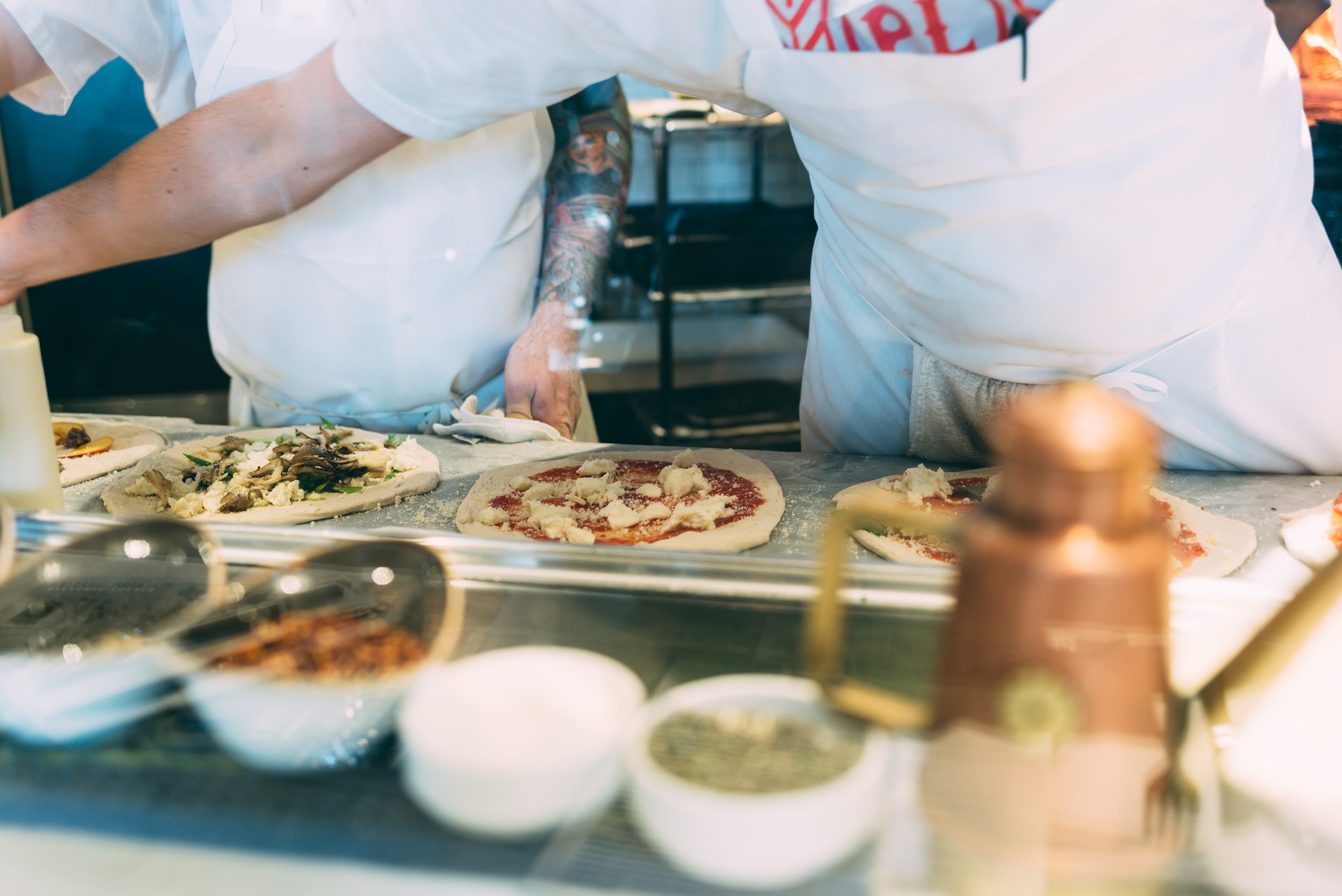 Chefs in white aprons assembling artisanal pizzas with mushrooms and cheese on a marble counter.