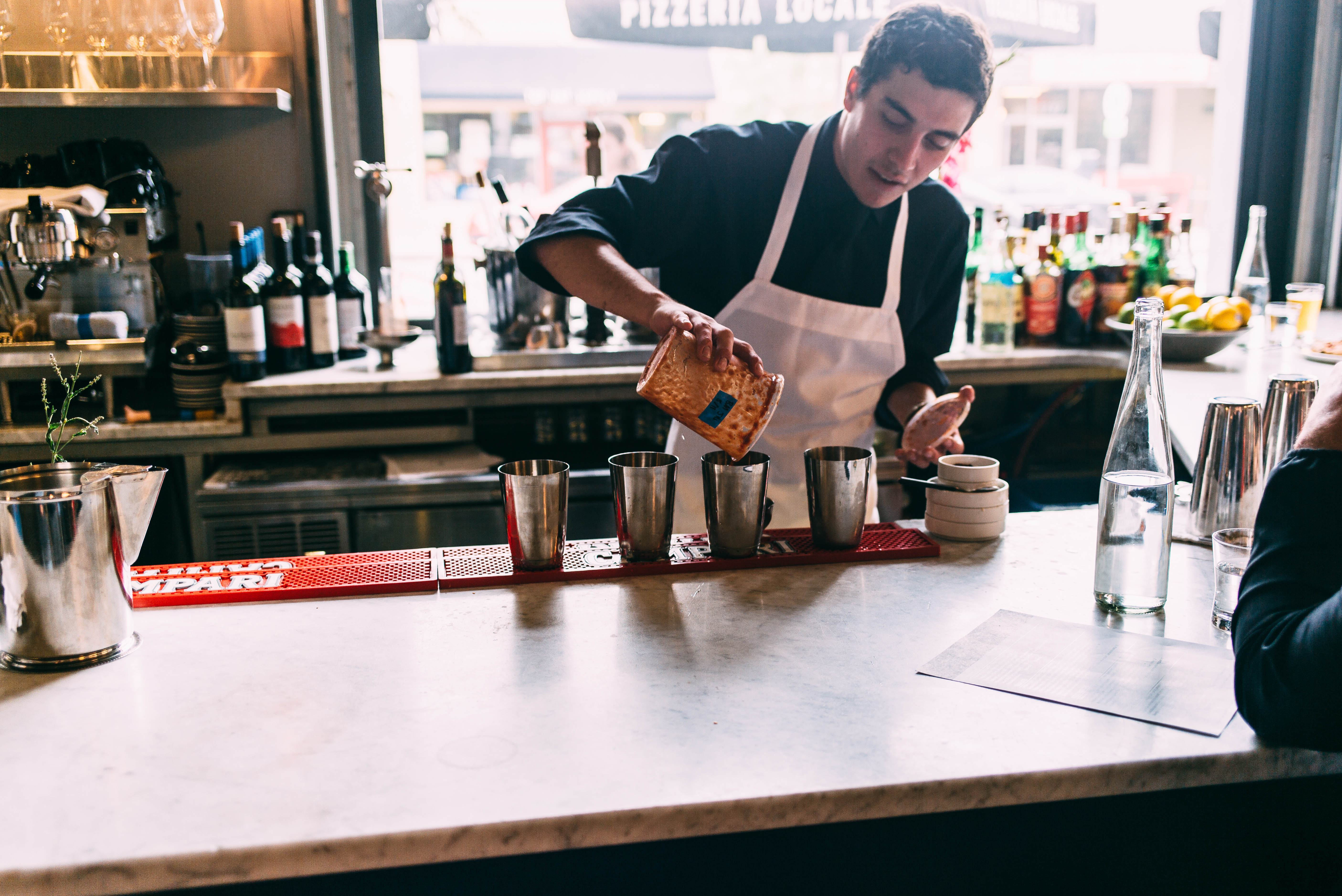 Bartender in an apron preparing cocktails behind a marble bar, pouring ingredients into shaker tins.