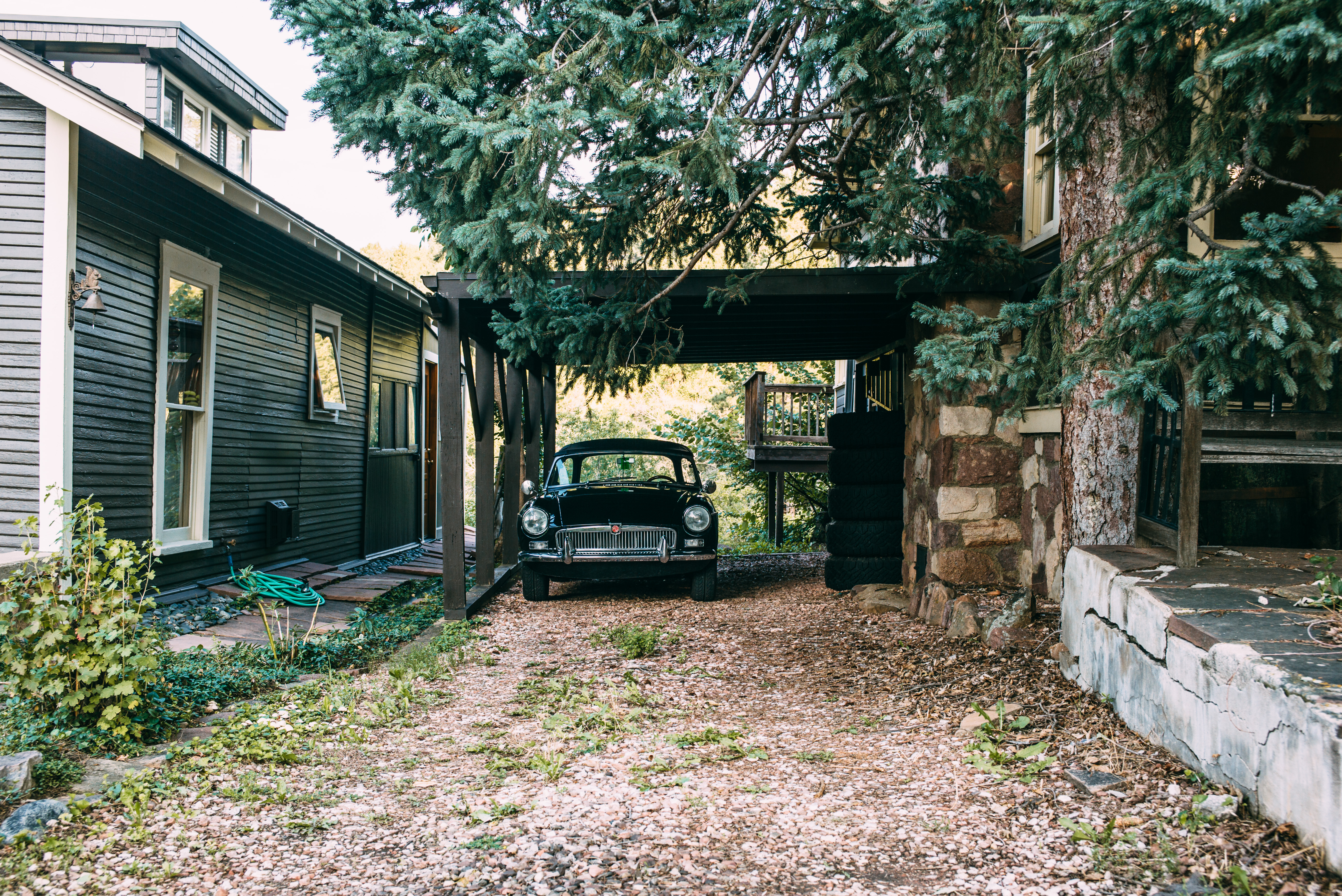 Black vintage MG roadster parked under a wooden carport beside a stone mountain home in Boulder.