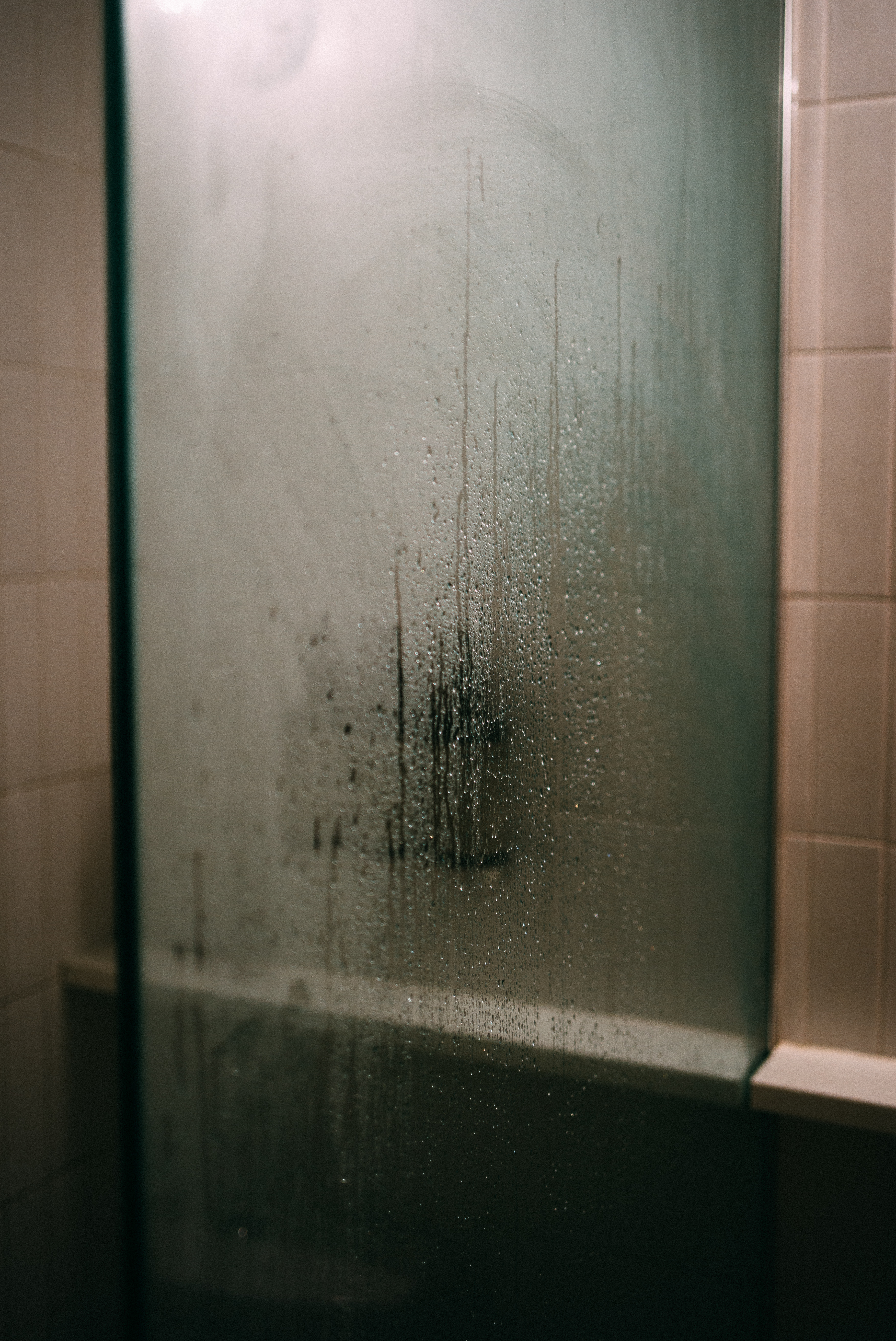 Water droplets and condensation streaks running down a steamy glass shower door in a tiled bathroom.
