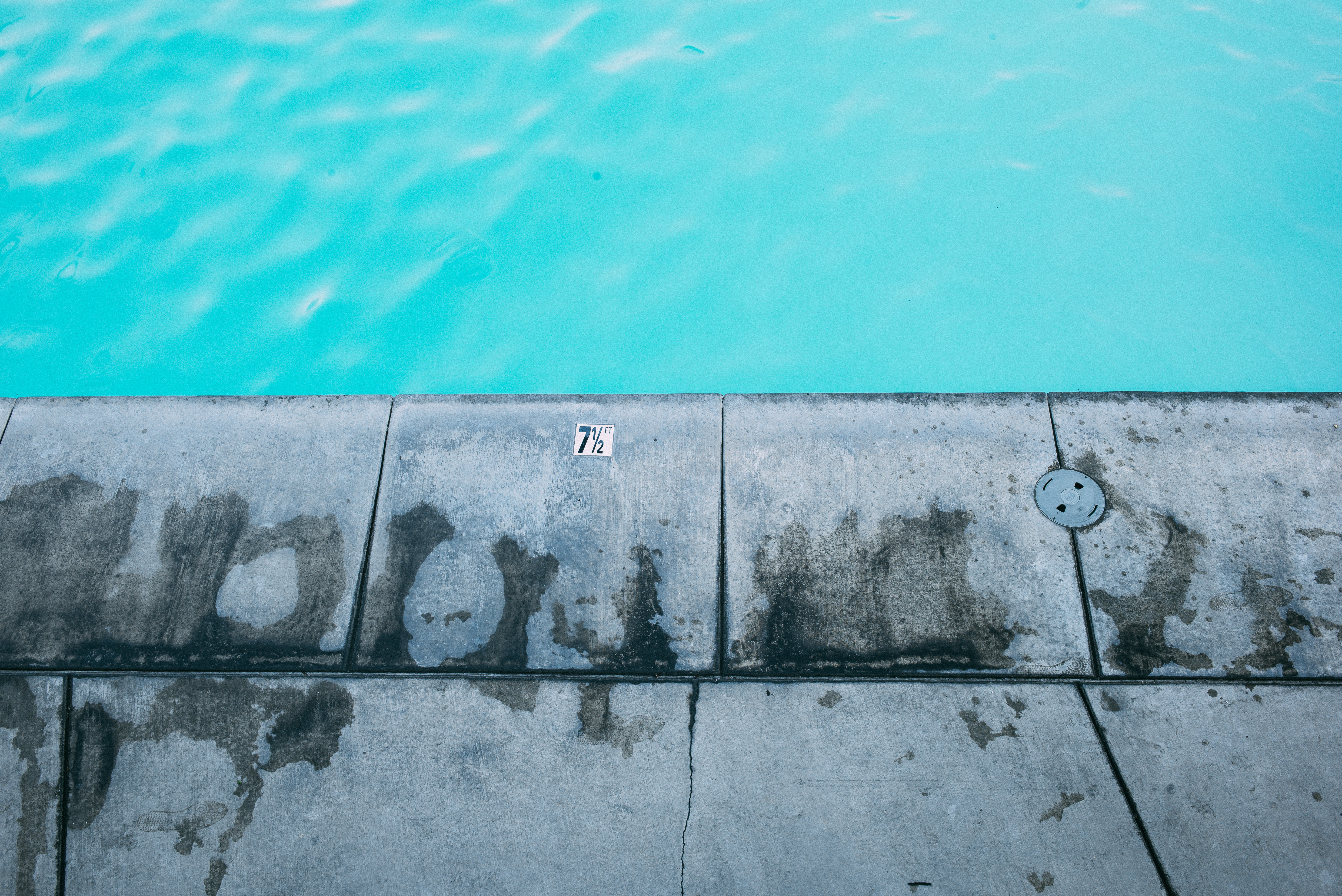 Depth marker tile reading '7 1/2 FT' on wet concrete coping with footprints beside turquoise pool water.