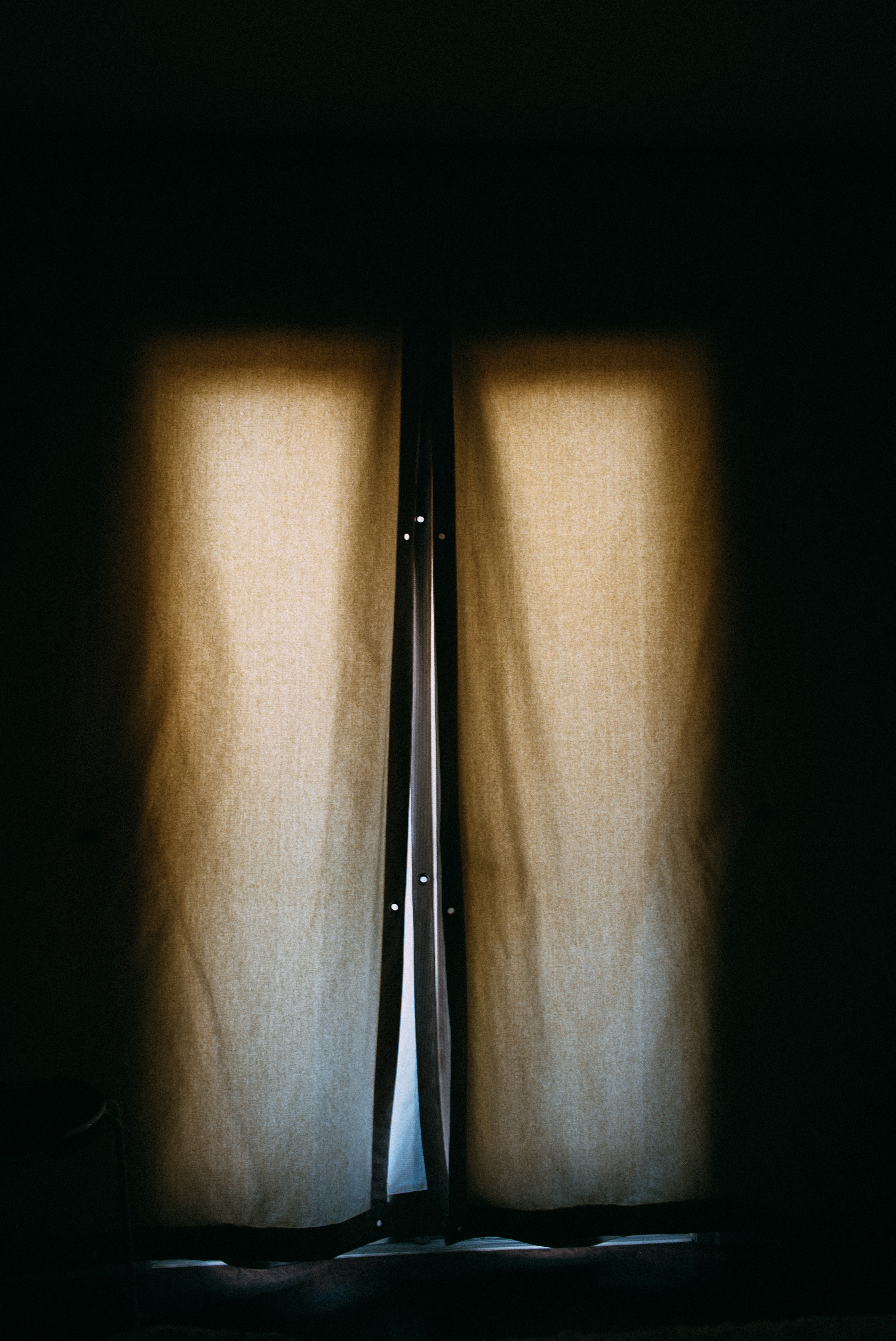 Sunlight glowing through woven beige curtains with light spilling through a narrow center slit in a dark room.