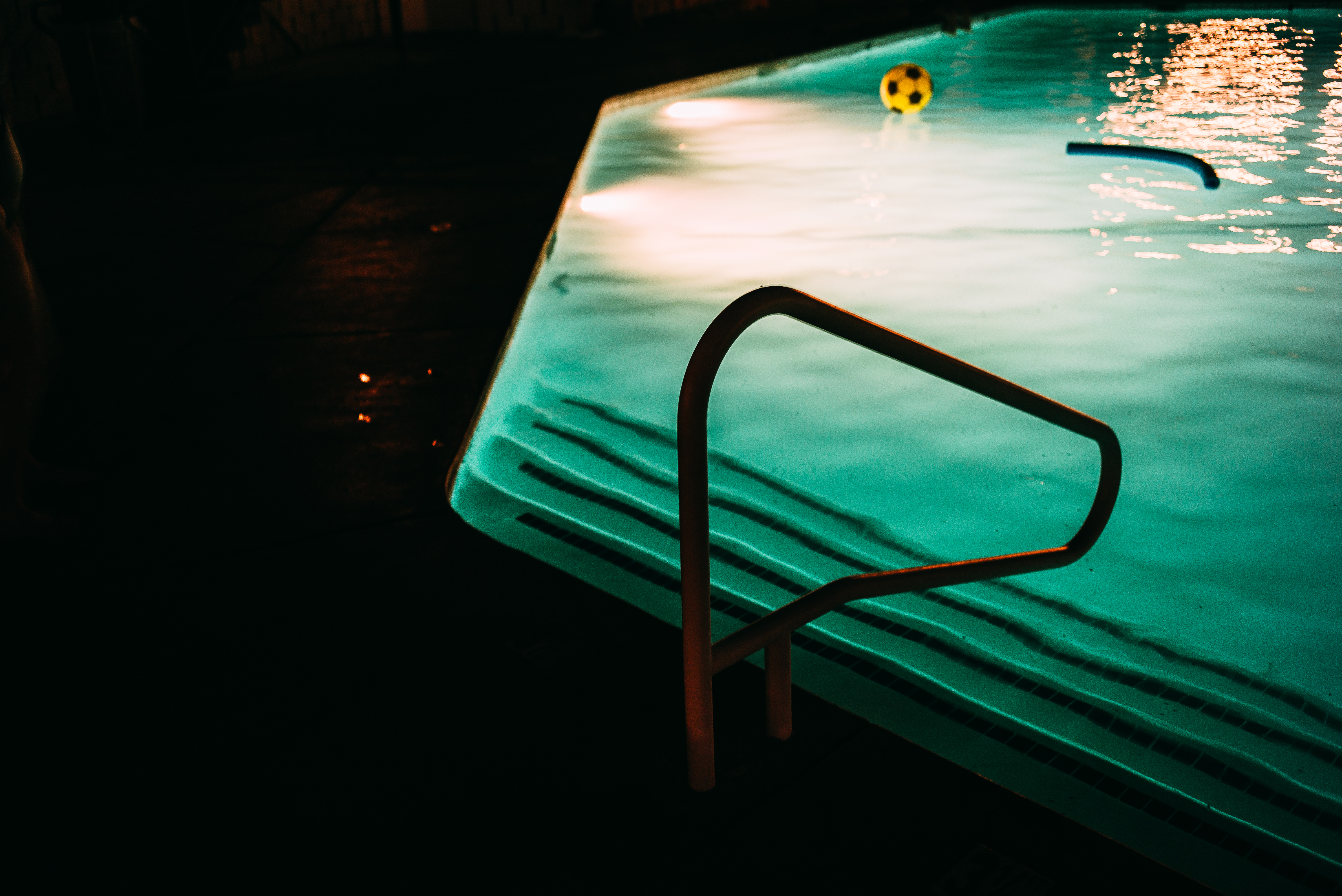 Illuminated turquoise swimming pool at night with a metal handrail and floating yellow soccer ball.