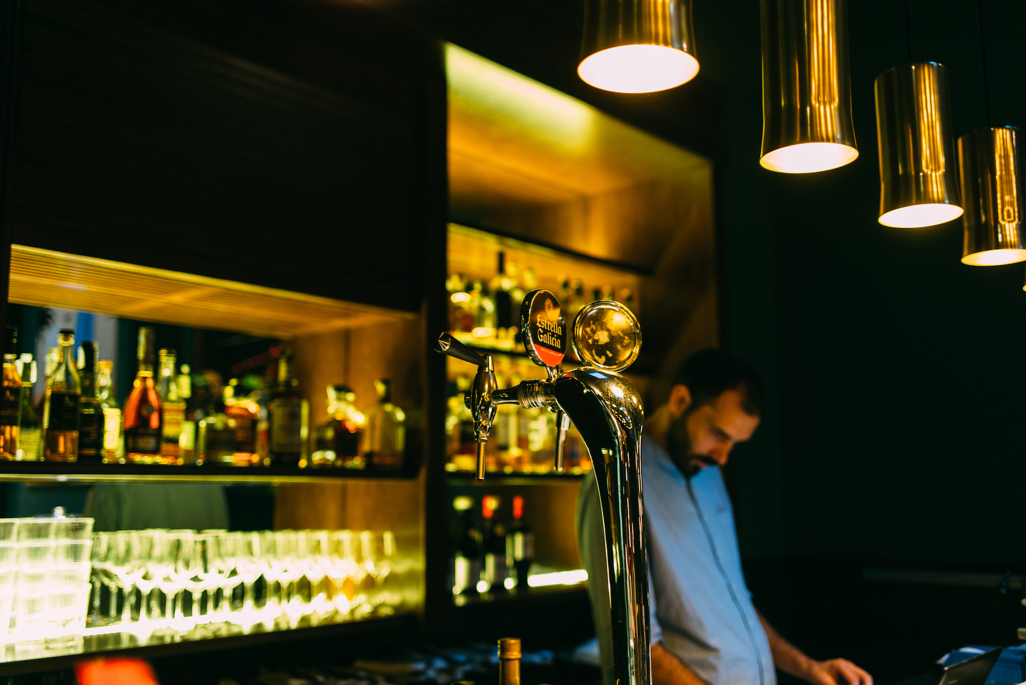 Chrome Estrella Galicia draft beer tap on a dark cocktail bar under warm brass pendant lights in Barcelona.