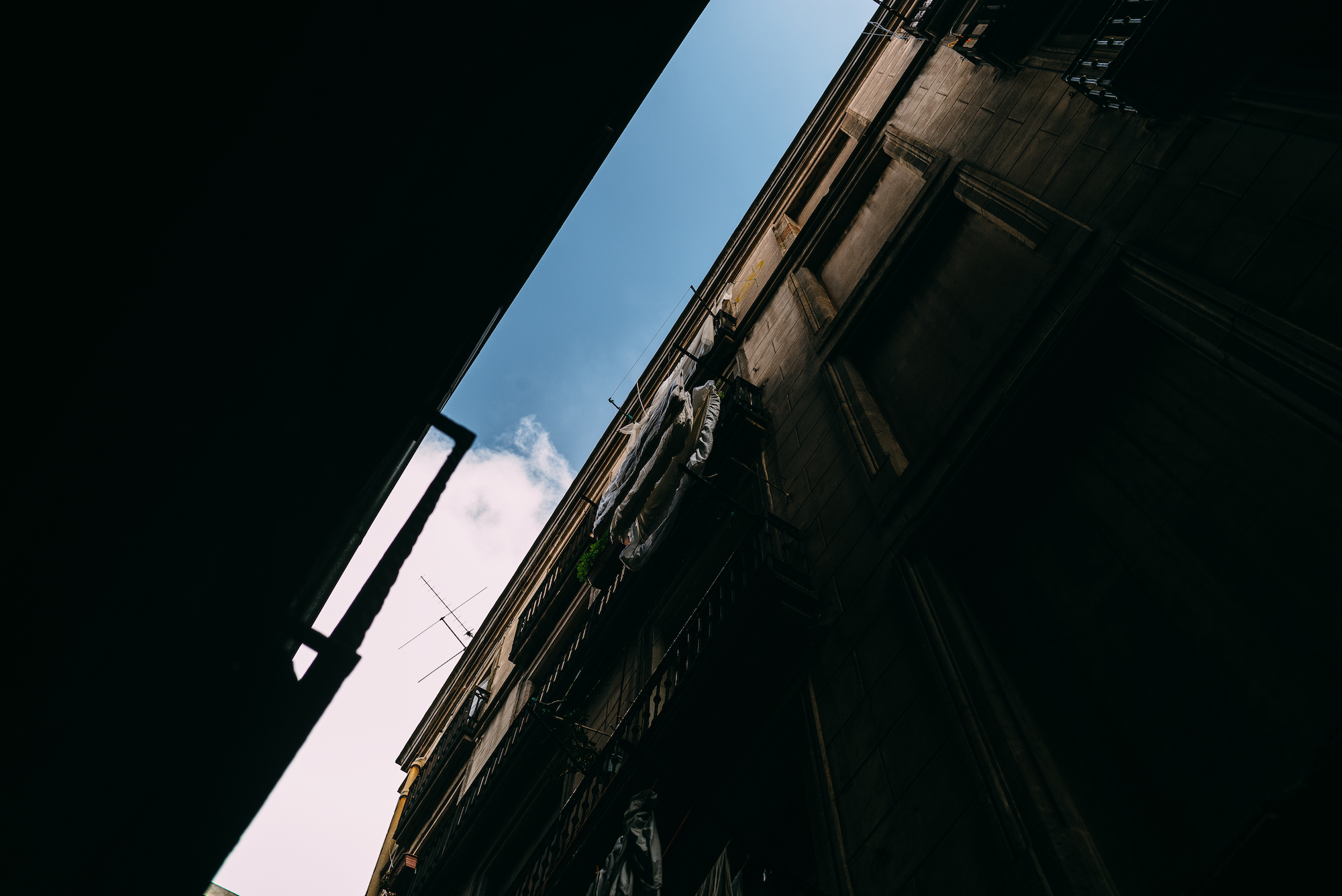 Dramatic upward-angle perspective looking between towering dark apartment facades and balconies toward a strip of bright blue sky in Barcelona, shot with a Leica M240.