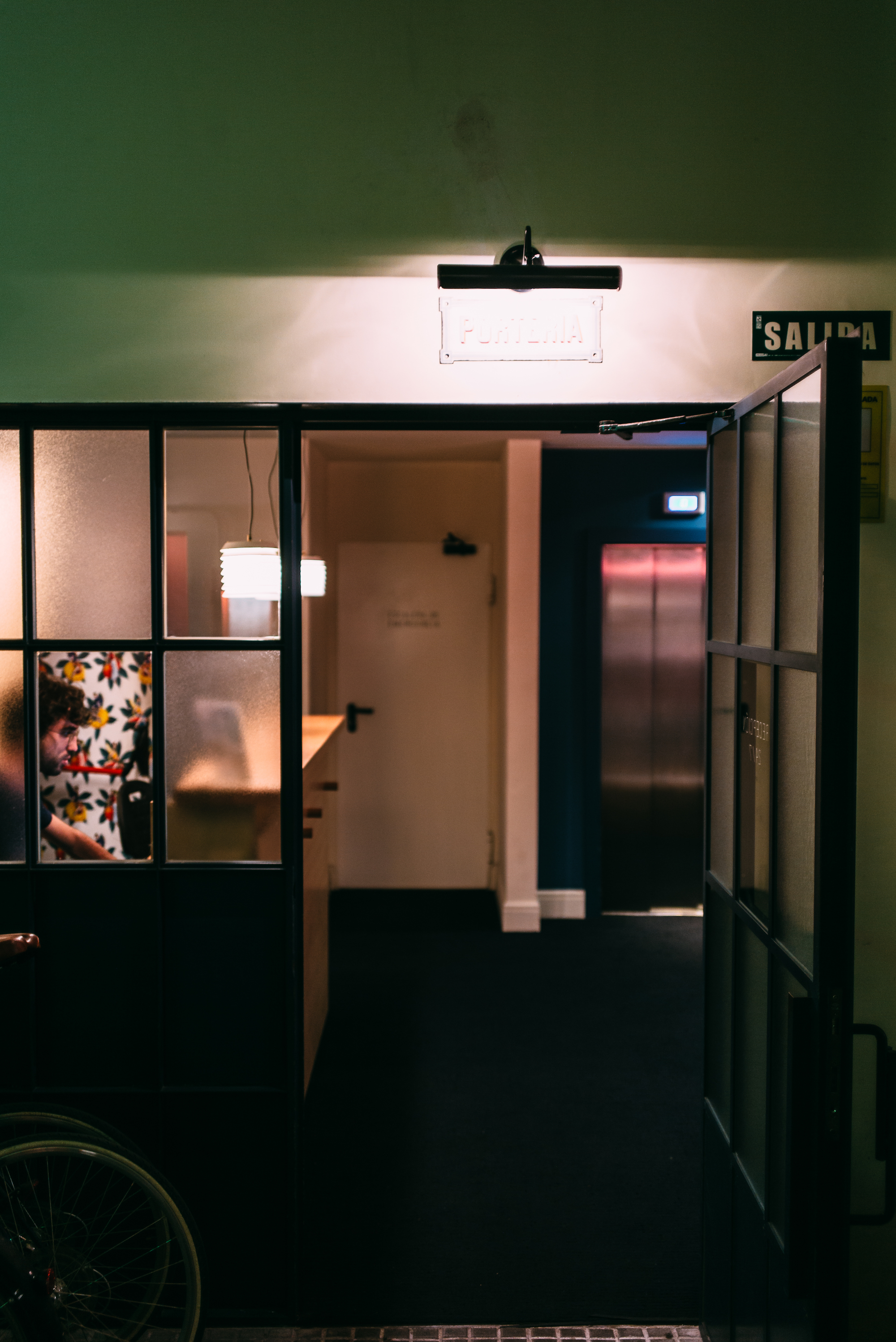 View through an open industrial glass frame door into a modern building entrance with a lit 'Porteria' sign, reception desk, and elevator lobby in Barcelona, shot with a Leica M240.