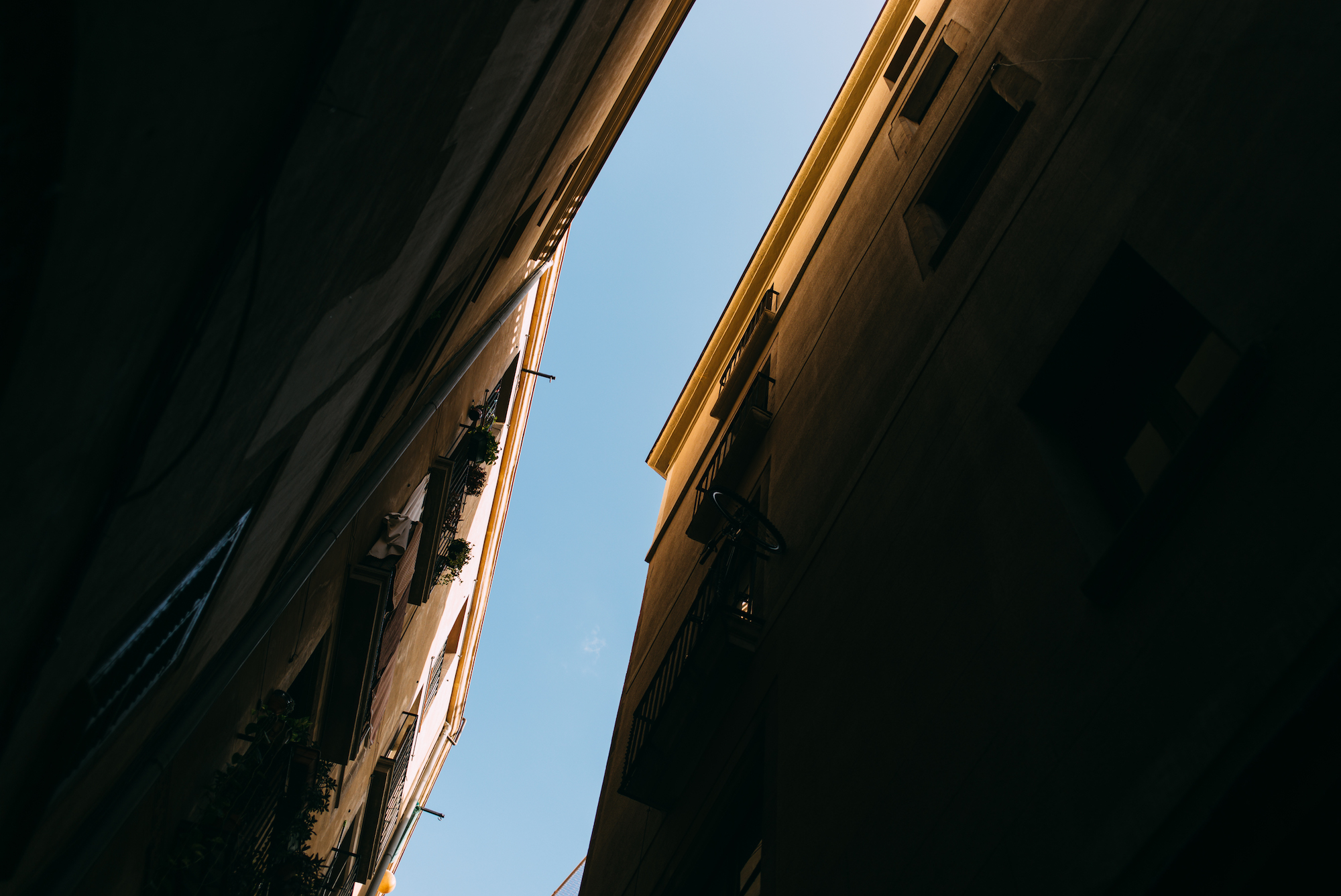 Geometric low-angle view looking upward between closely spaced apartment facades toward a sharp triangular strip of open sky in Barcelona, shot with a Leica M240.