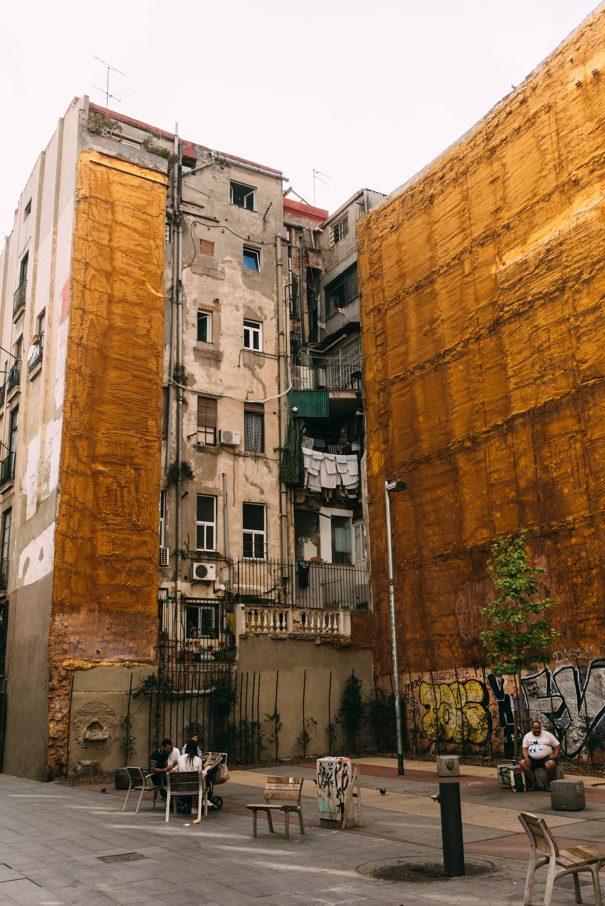 People seated on benches in an open urban plaza surrounded by weathered multi-story apartment buildings with vibrant yellow insulation walls and graffiti in Barcelona, shot with a Leica M240.