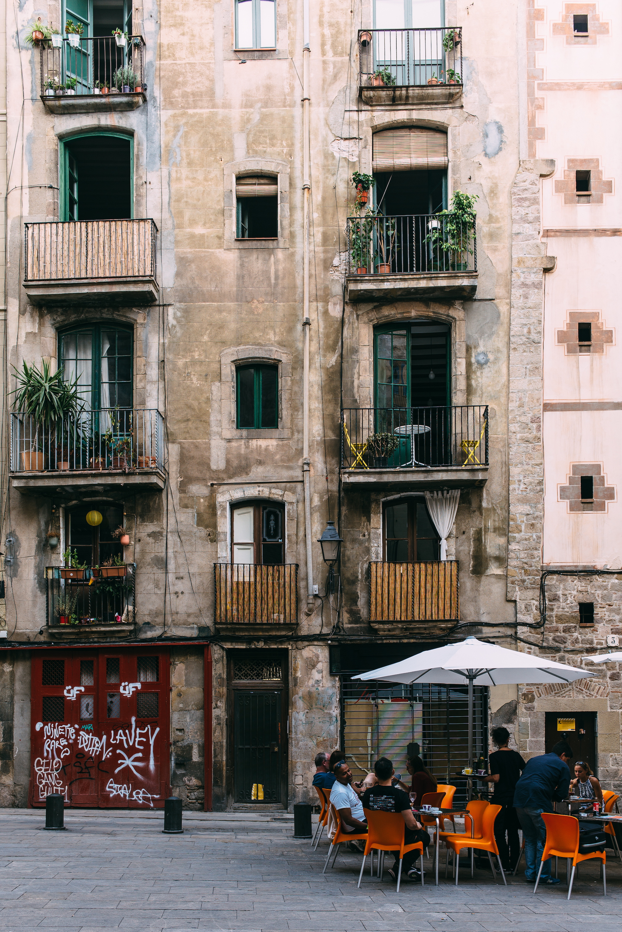 Patrons dining at orange patio chairs beneath a sun umbrella outside an aged apartment facade with balconies full of potted plants in a Barcelona square, shot with a Leica M240.