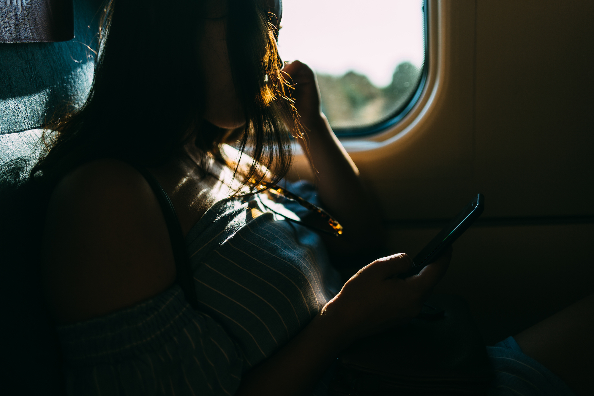 Silhouetted woman on a Renfe train looks at her phone with sunlight highlighting her hair and shoulder.
