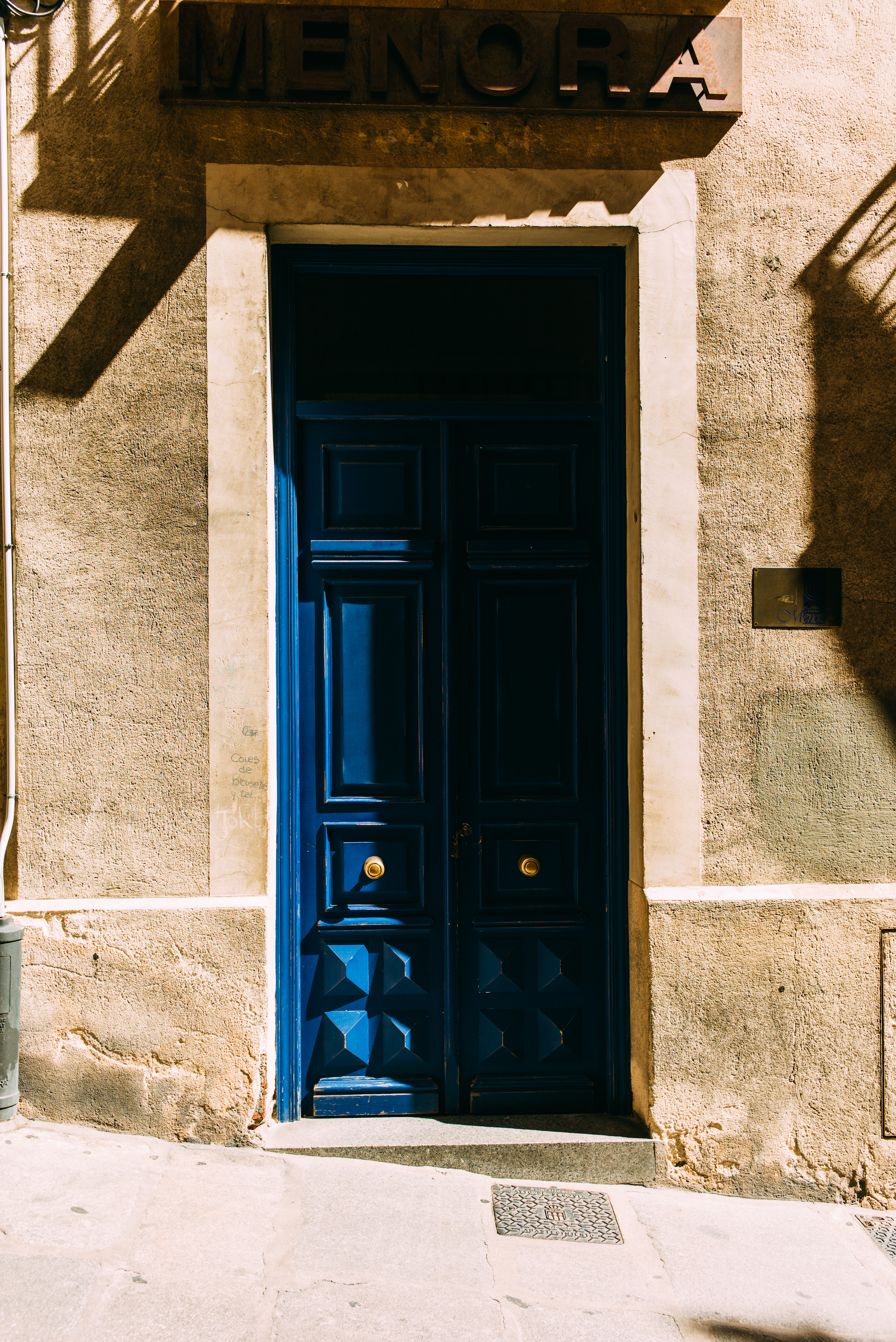 Blue wooden door set in a beige stone facade under a rusty metal "MENORA" sign on a Madrid street.