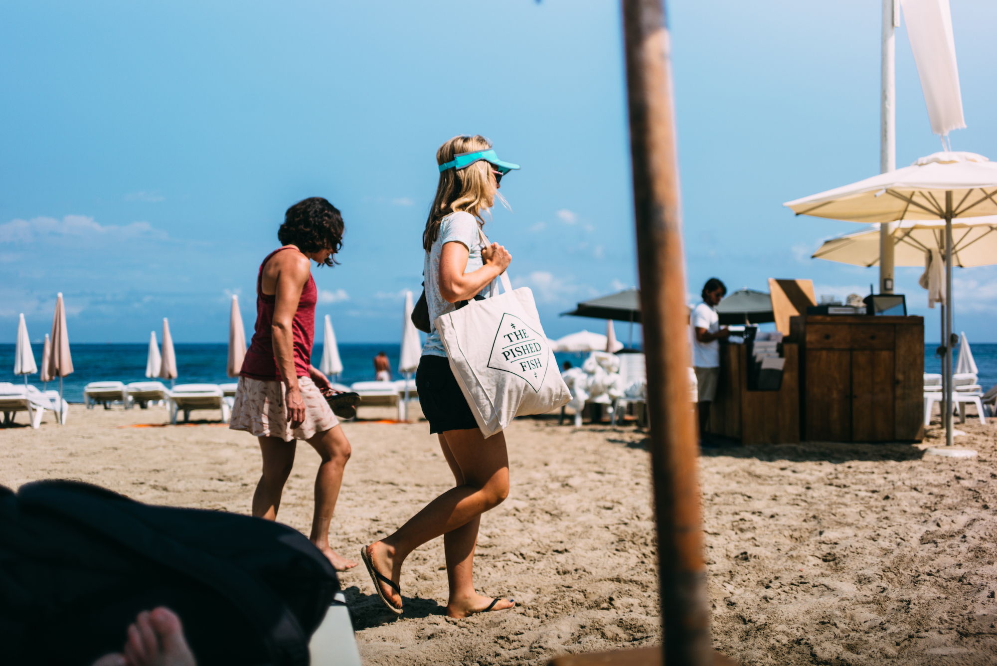 Two women walk past beach umbrellas, one carrying a 'The Pished Fish' tote bag in Formentera.