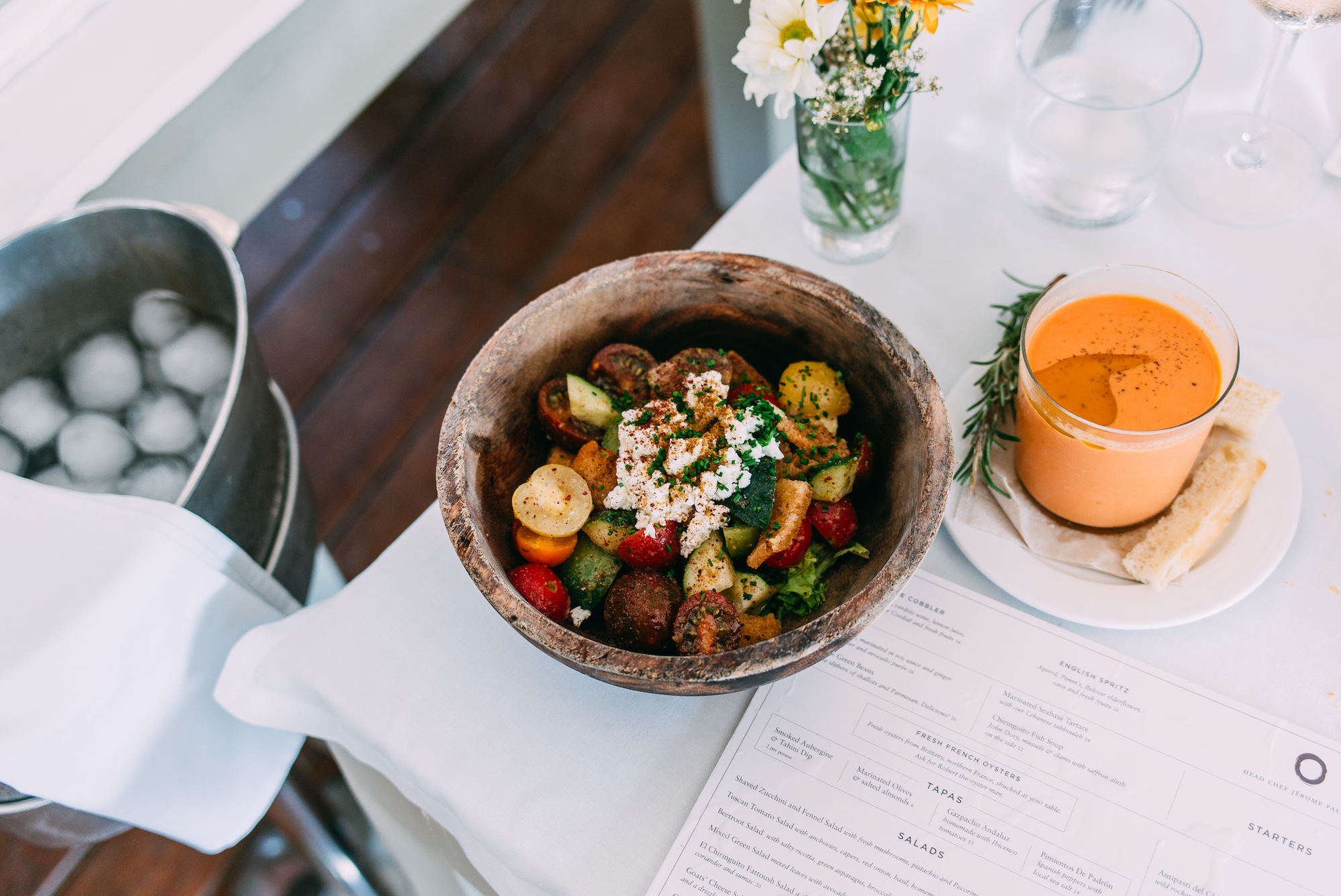 Panzanella salad in a wooden bowl served beside chilled gazpacho and a cocktail menu on an outdoor table.