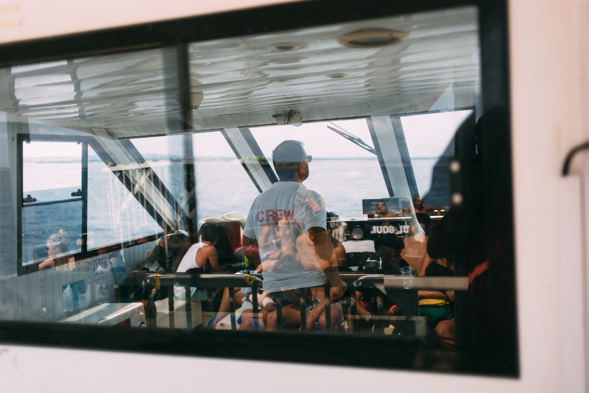 Ferry crew member in a 'CREW www.capitanhook.es' shirt looking out to sea while passengers sit inside.