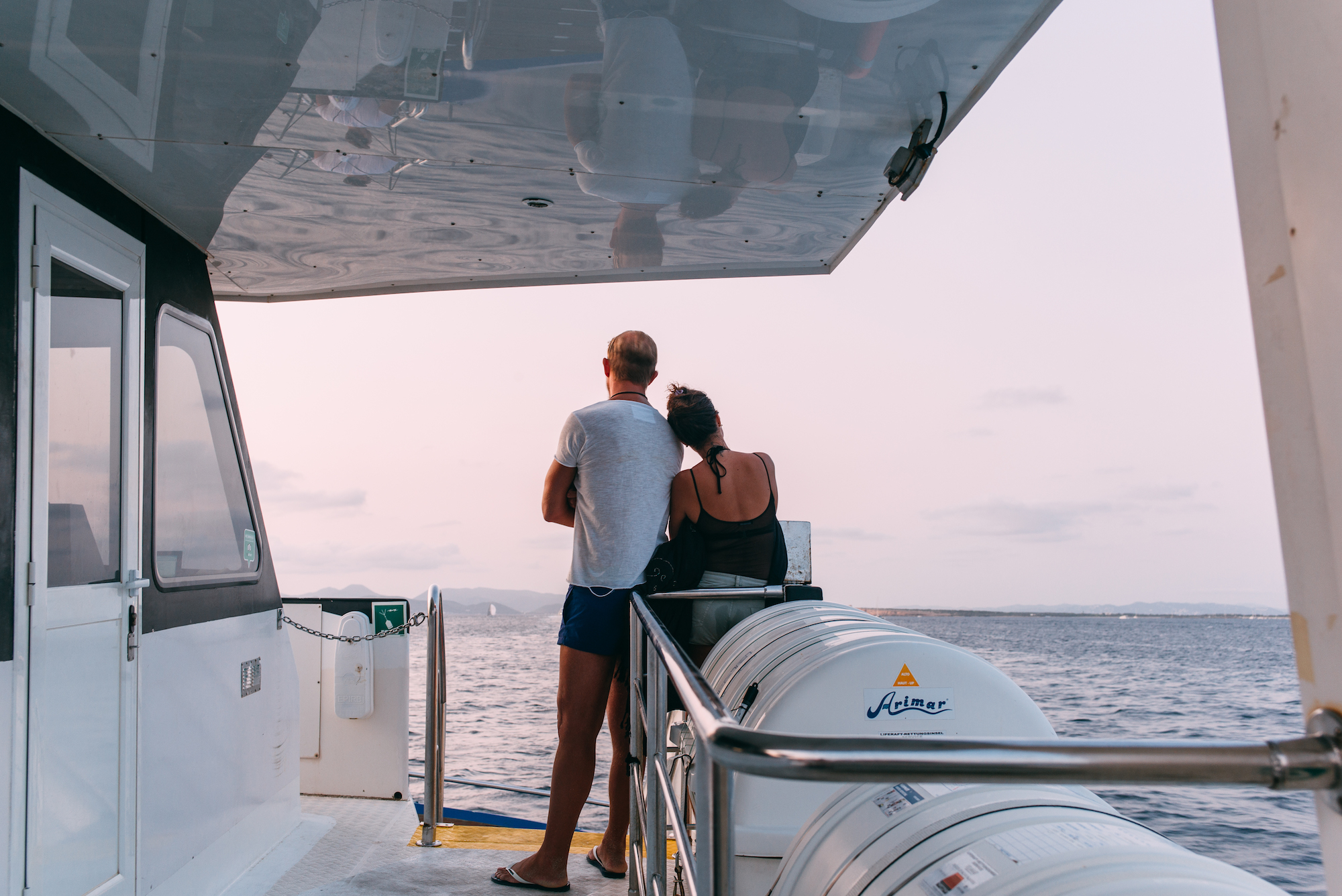Couple at the stern railing of a ferry boat looking out across the sea toward coastal hills at sunset.