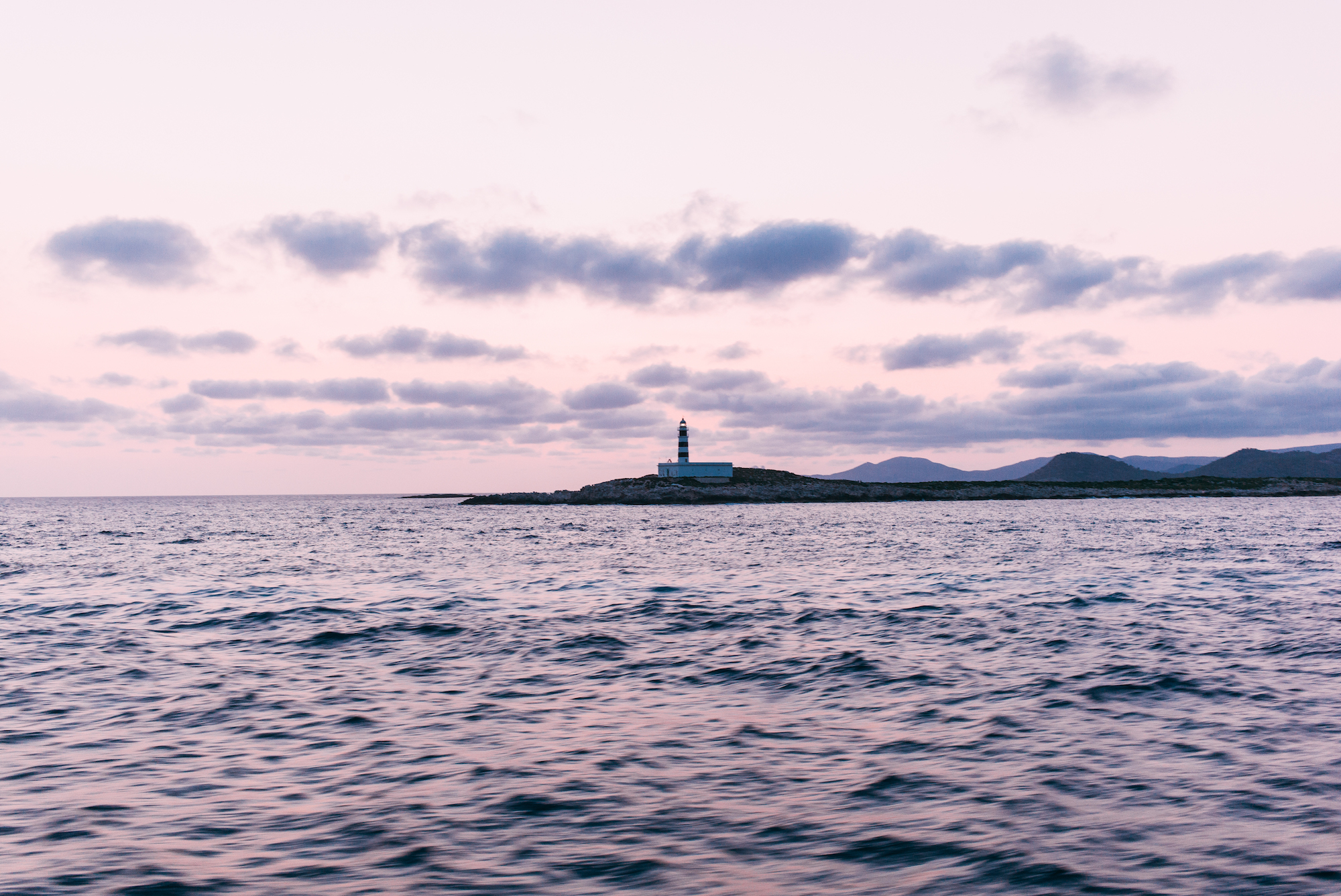 Black and white striped lighthouse on a rocky island across dark sea waters under sunset clouds.