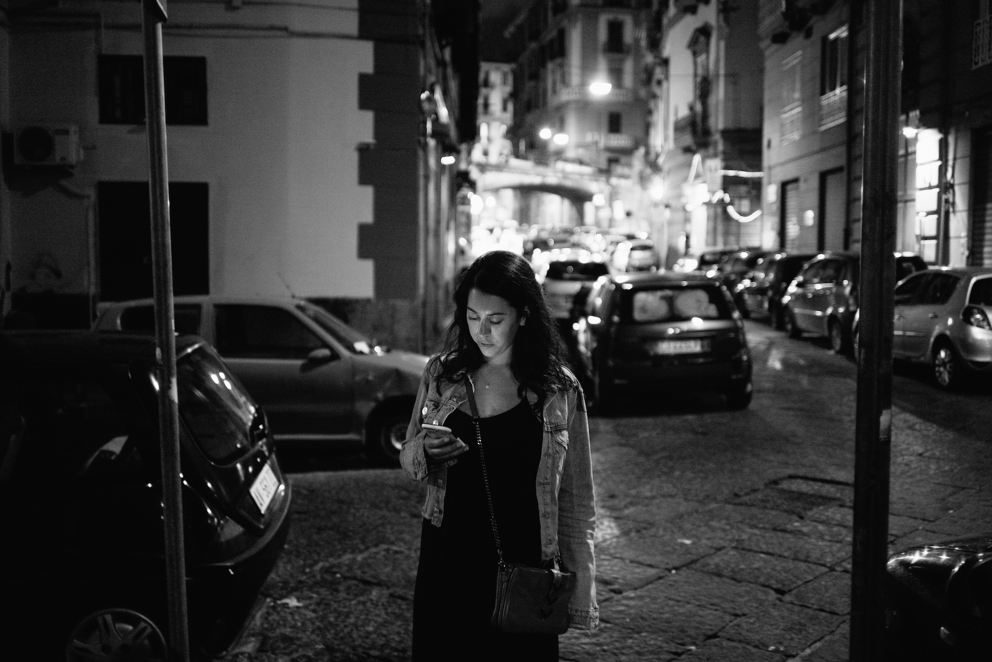Young woman in a denim jacket looking at her smartphone on a cobblestone city street lined with cars at night.