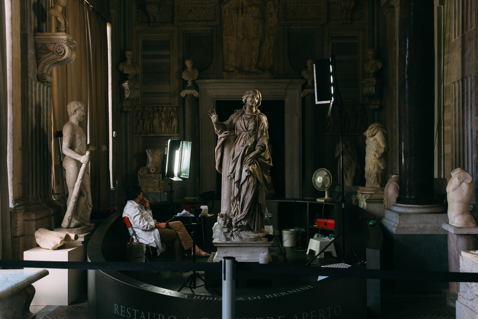 Conservator in a white coat illuminating Bernini's marble statue of Santa Bibiana with studio lights in Rome.