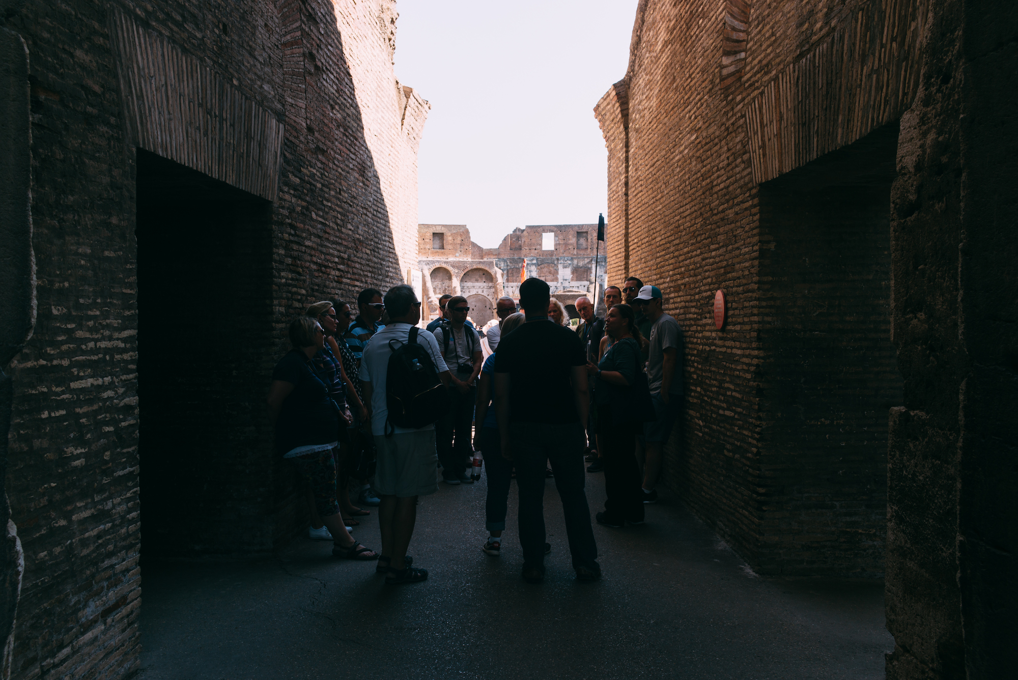 Tour group in a shadowed Roman brick passageway looking toward the sunlit arches of the Colosseum in Rome.