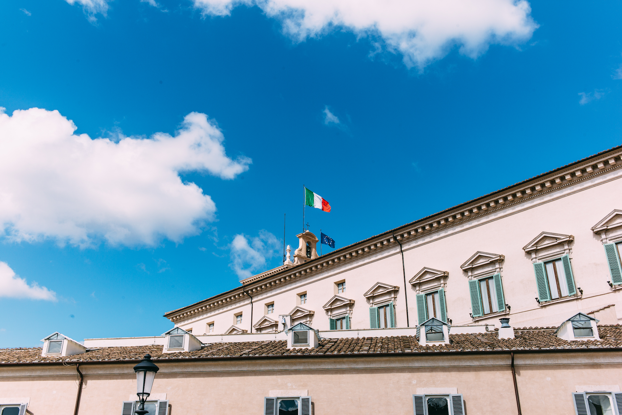 Italian and EU flags fluttering on the Quirinal Palace rooftop bell tower under a blue sky in Rome.