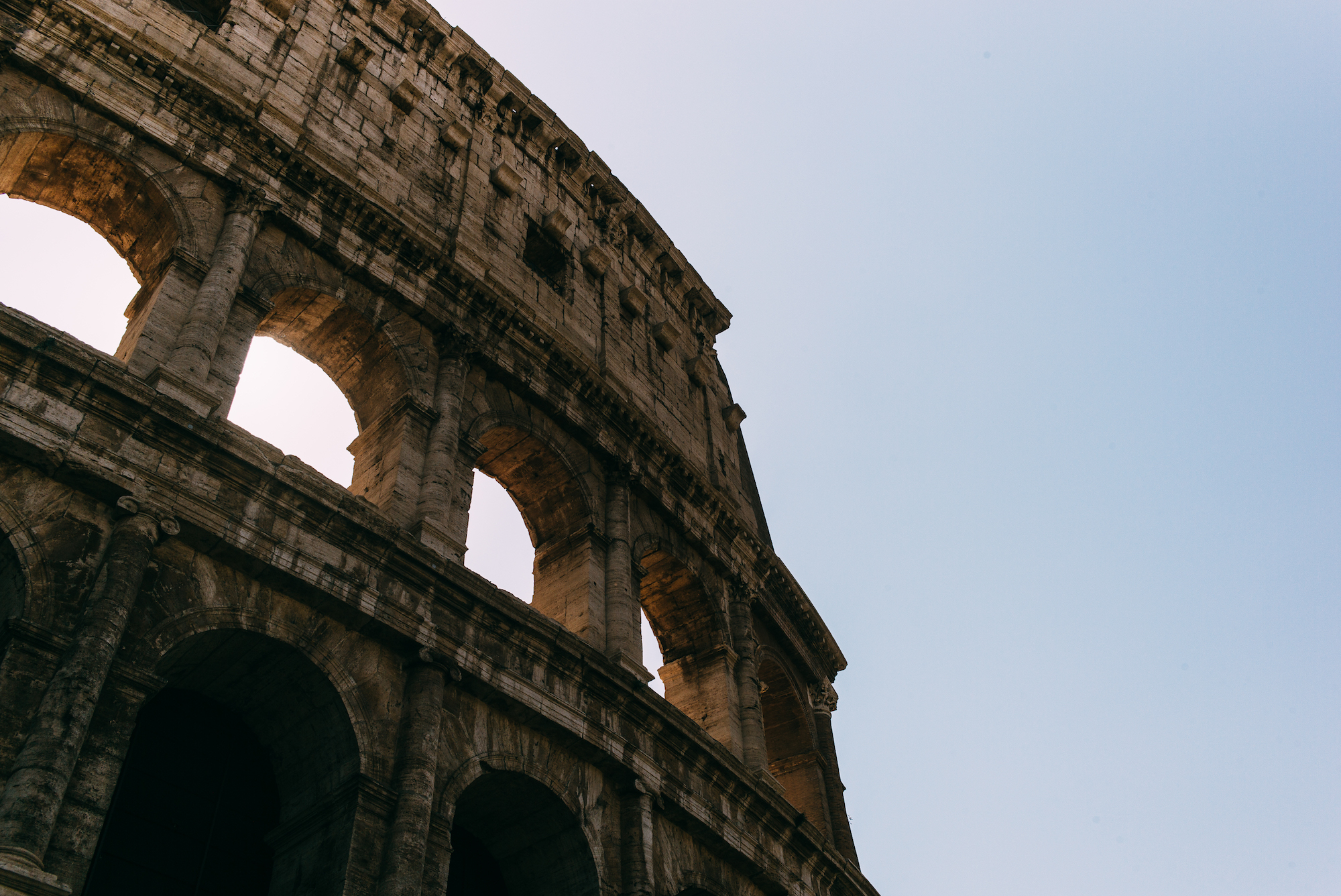 Exterior stone arched tiers of the Colosseum bathed in warm afternoon light against a blue sky in Rome.