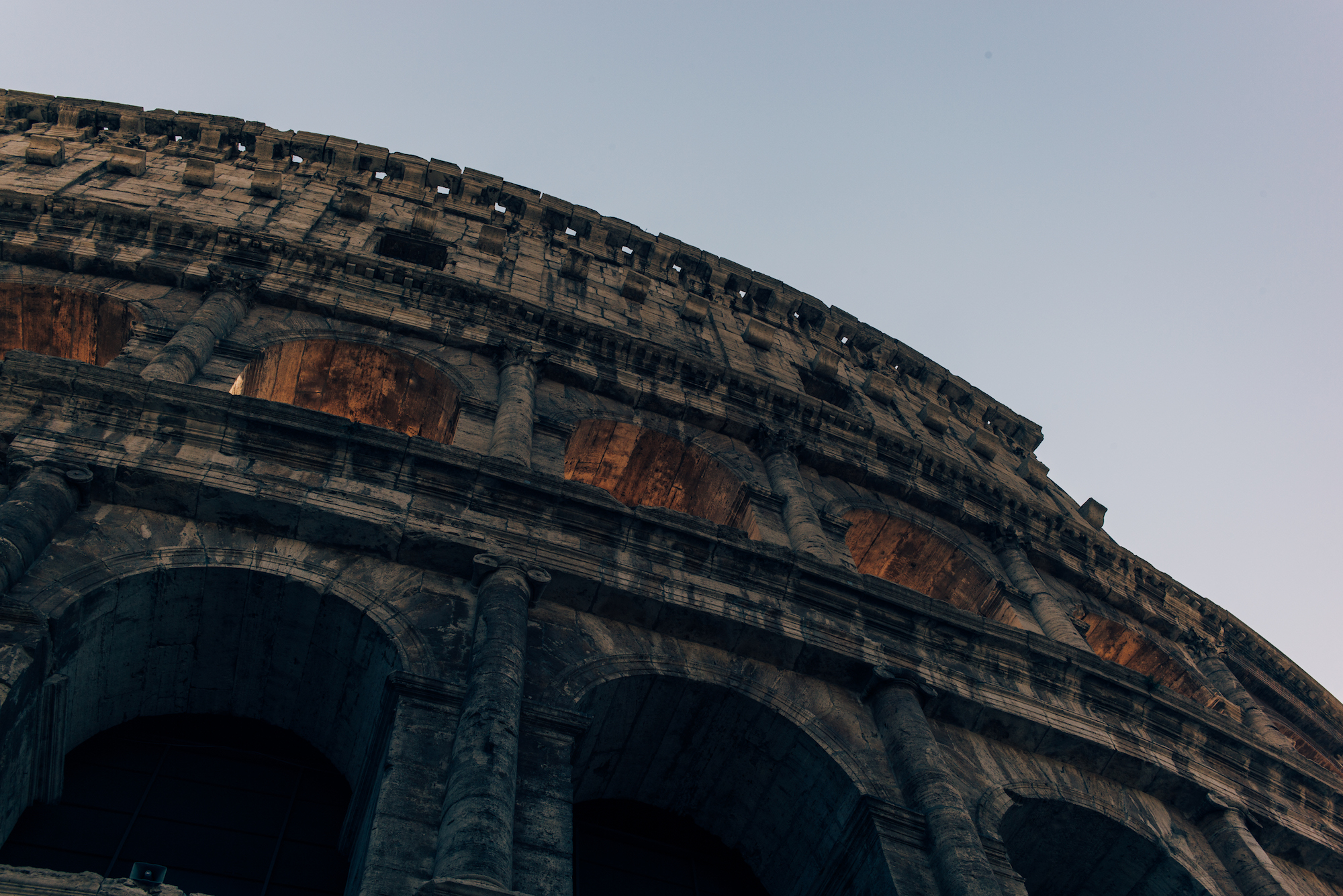 Monumental travertine arches and upper attic level of the Colosseum against an evening sky in Rome.