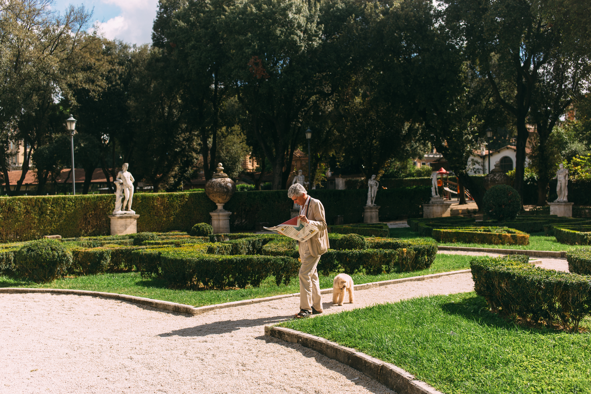 Italian gentleman in a beige suit reading a paper while walking a dog in a formal garden in Rome.