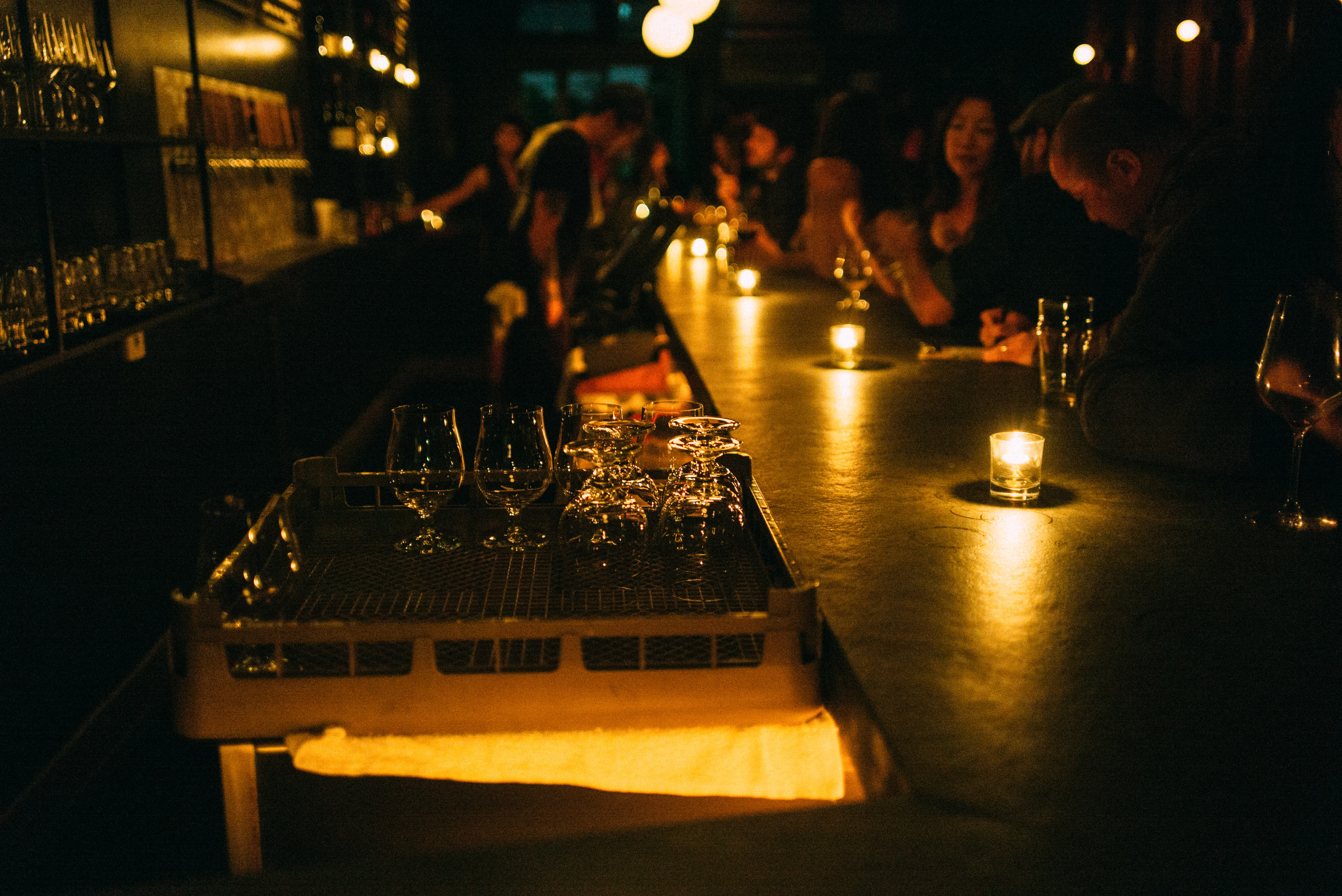 Dimly lit bar counter in warm candlelight with votive candles reflecting off the surface as patrons socialize.