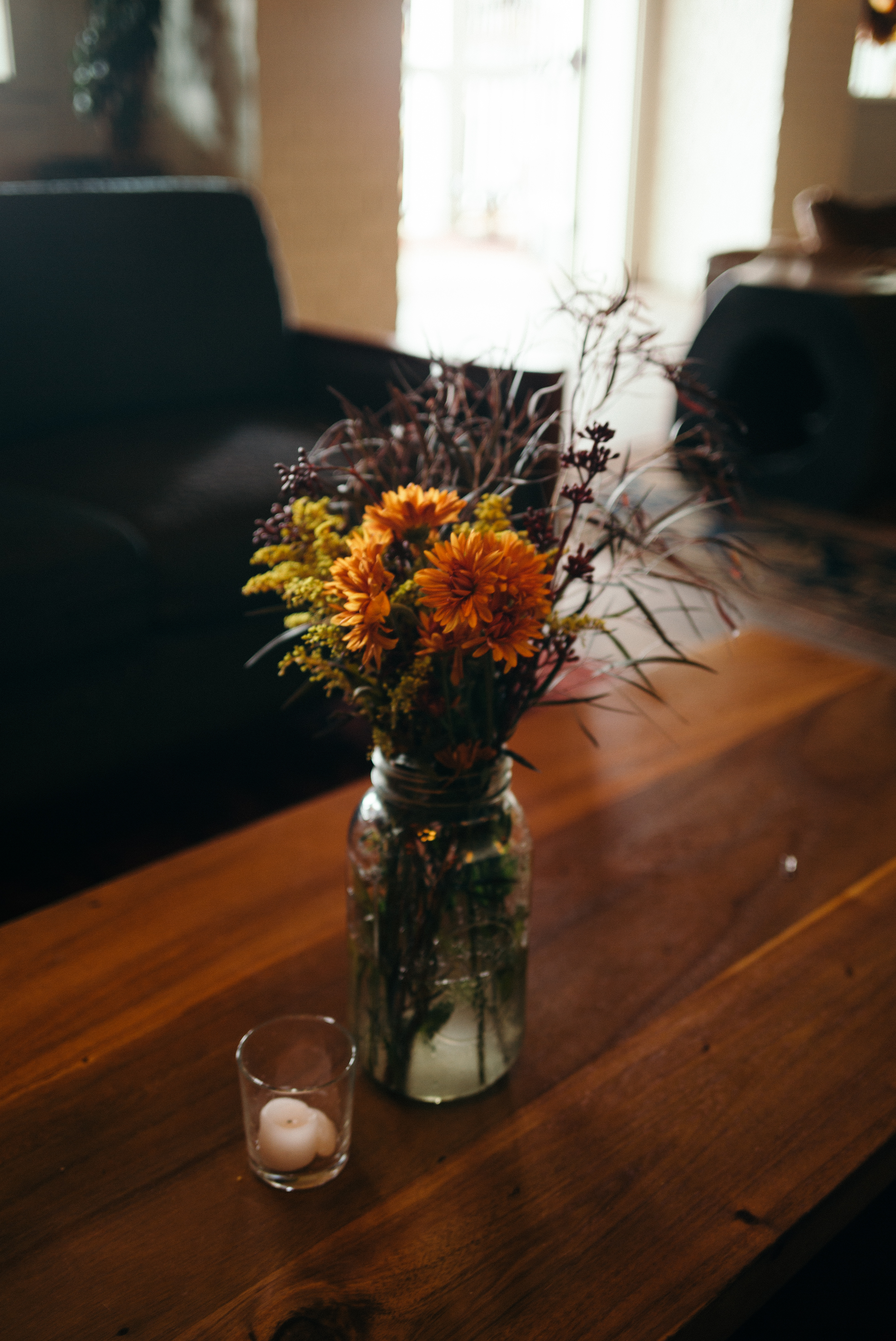 Glass mason jar of orange chrysanthemums and greenery beside a votive candle on a wood coffee table.