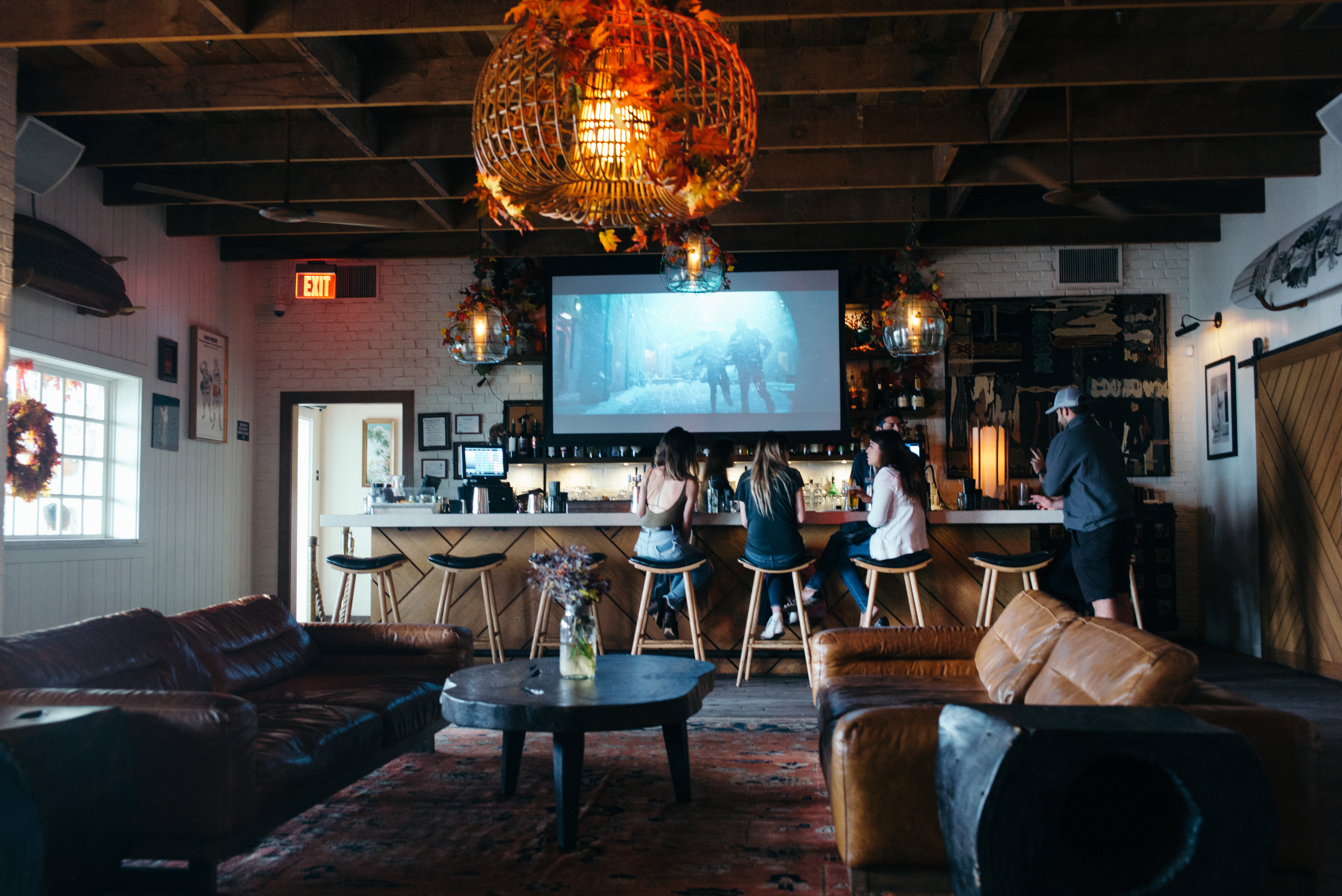 Lodge lounge with leather sofas and a wood-paneled bar where guests watch a screen under a wicker lamp.