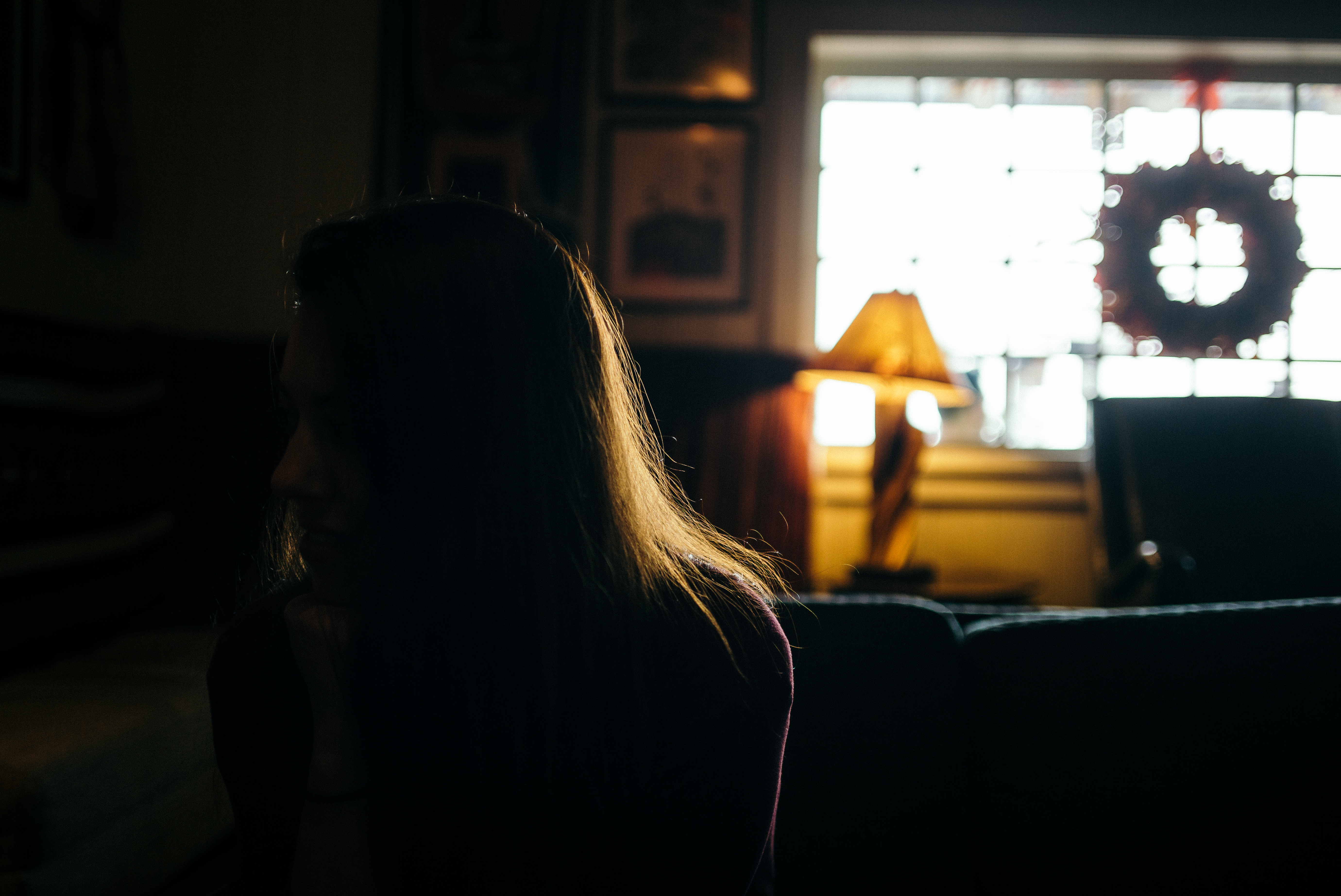 Backlit silhouette of a woman with rim-lit hair seated in a dim room beside a glowing table lamp.