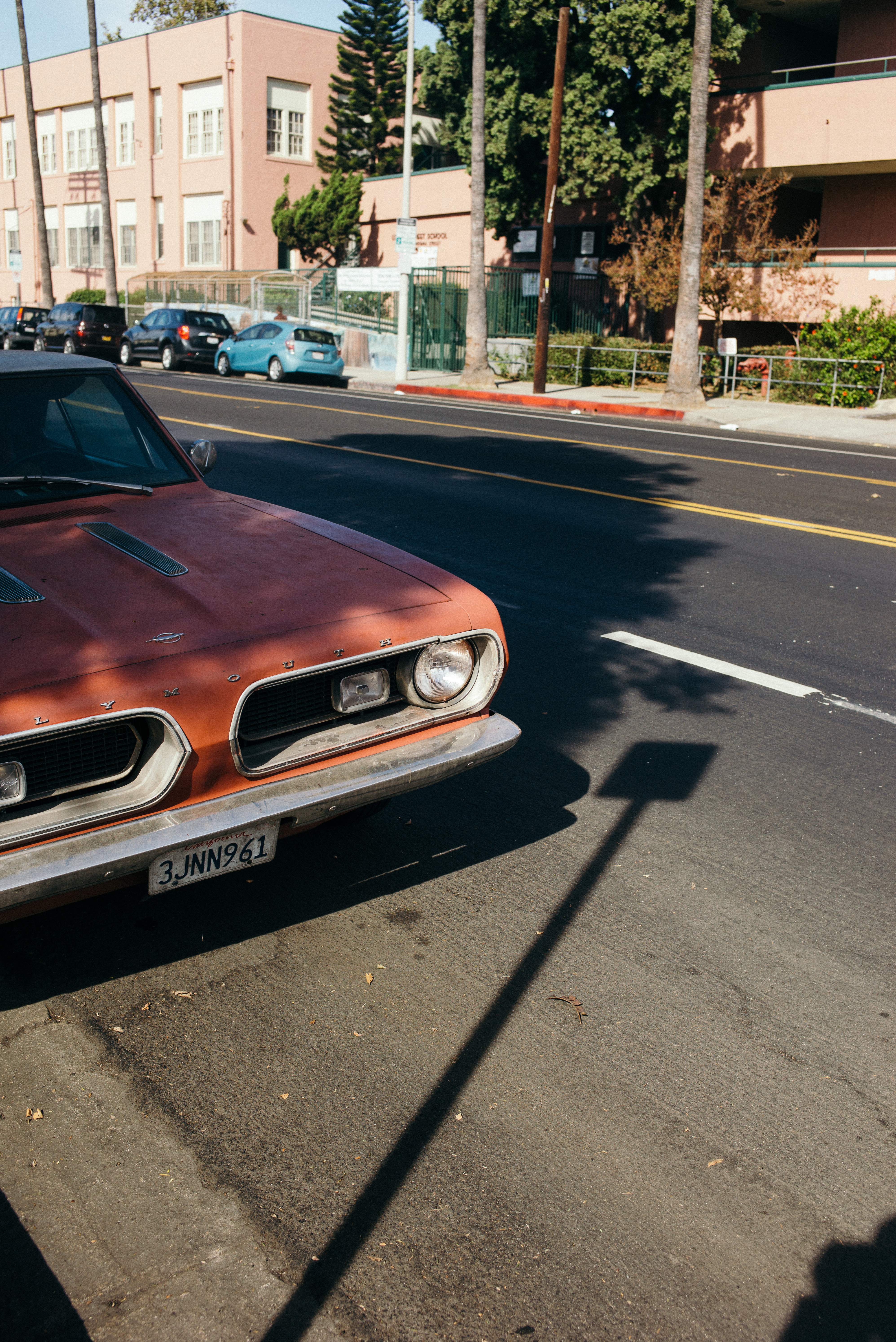Faded orange vintage Plymouth Barracuda parked along a sunny street with palm trees and stucco buildings.