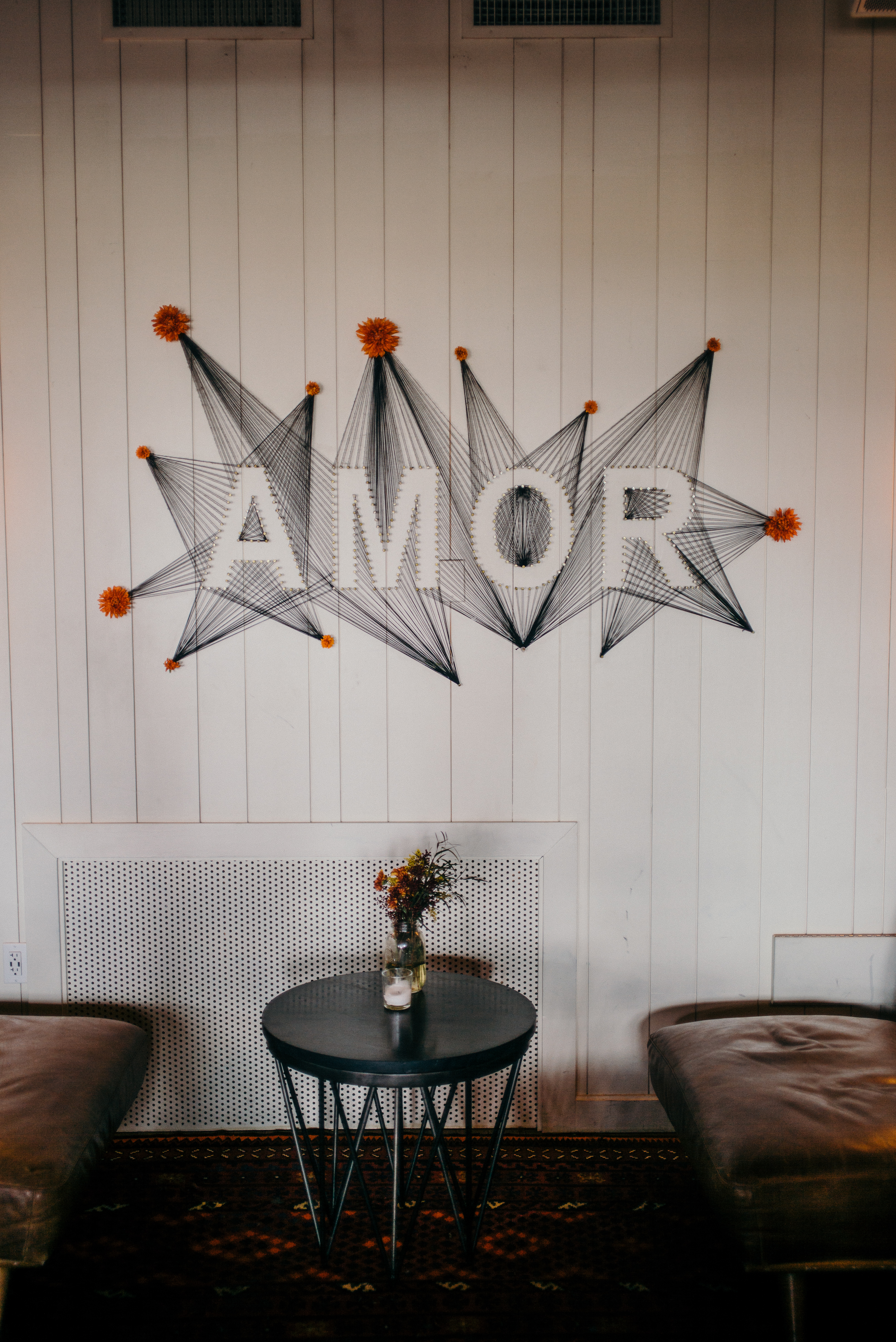 Wood-paneled wall with string art spelling 'AMOR' accented with orange flowers above leather chairs.