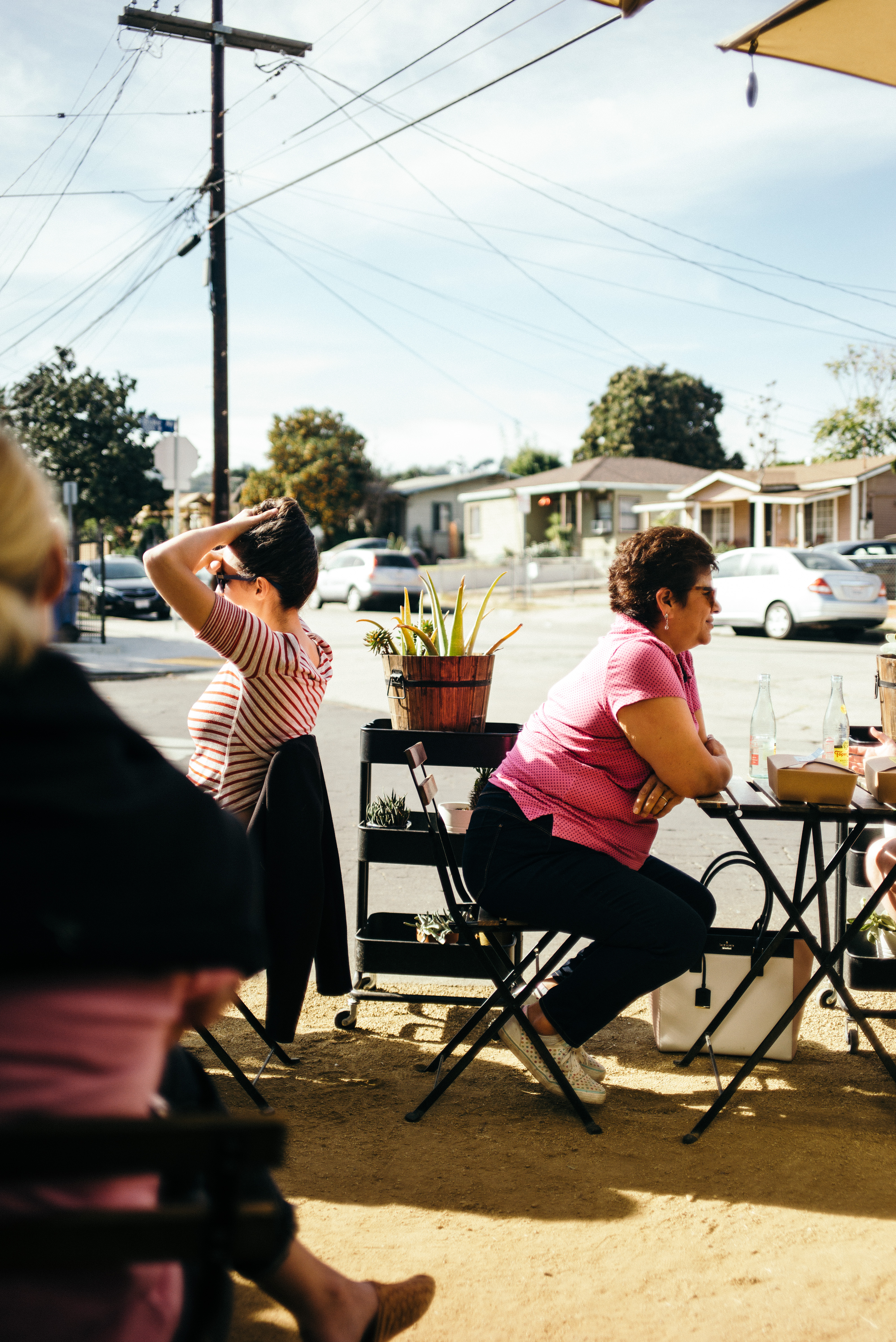 Two women seated at an outdoor cafe patio table on a sunny day in Los Angeles, one in a pink polo shirt and the other in a striped top adjusting her hair, shot with a Leica M240.
