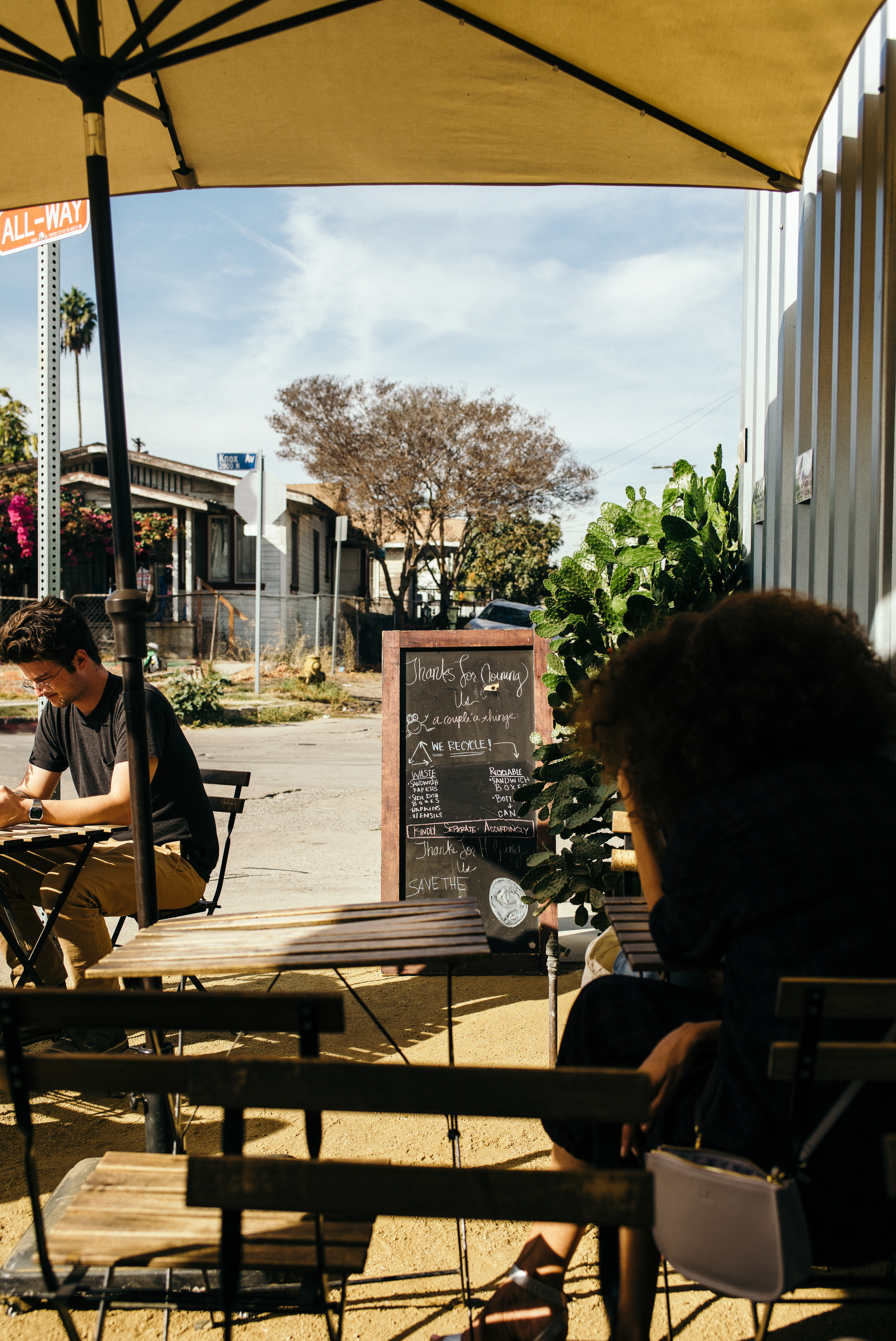 Patrons sitting under a yellow cafe umbrella at outdoor wooden tables beside a chalk blackboard menu reading 'Thanks for joining us' and a tall green prickly pear cactus in Los Angeles, shot with a Leica M240.