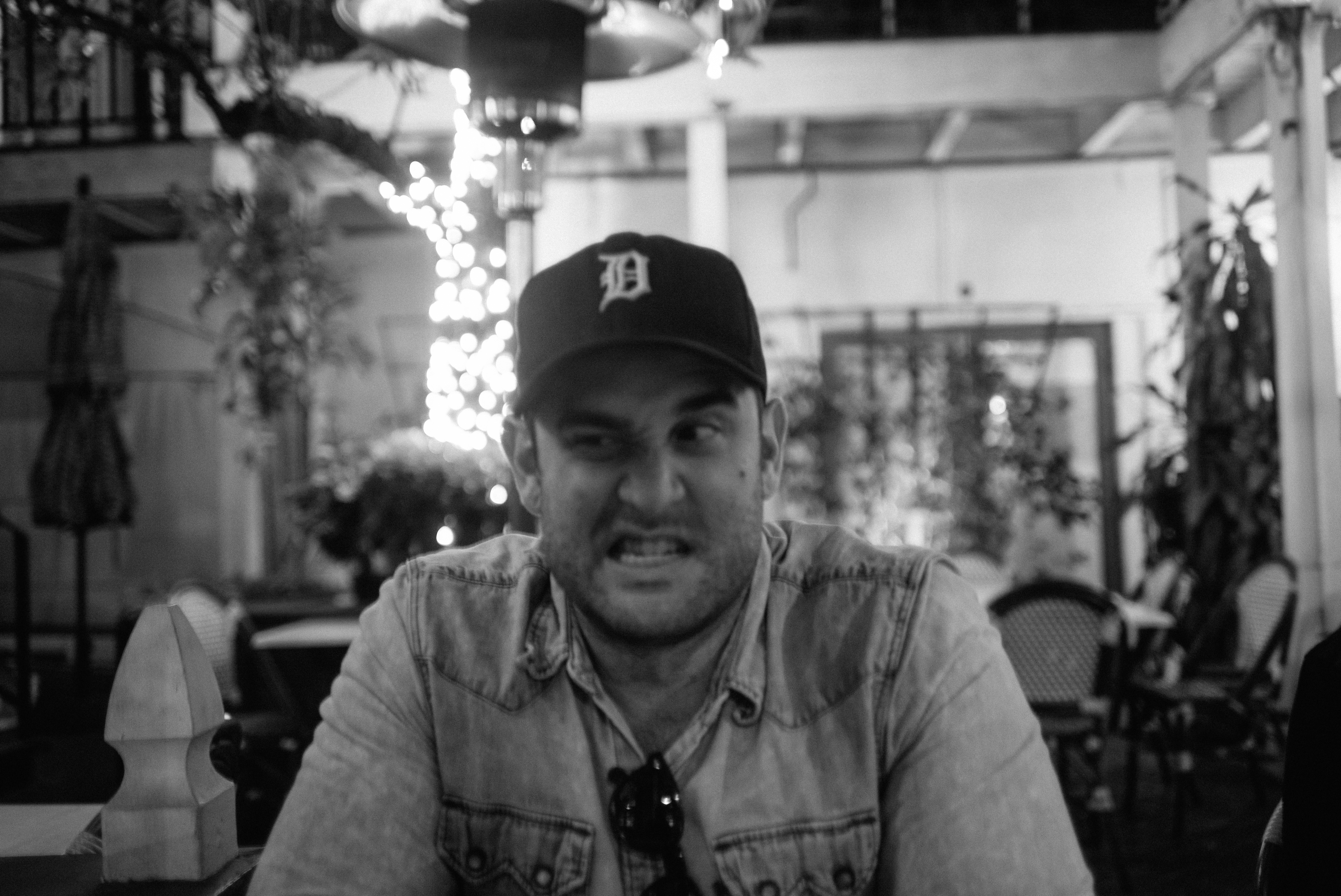 A black and white portrait of a man in a Detroit Tigers baseball cap and denim shirt making a playful grimacing face at an outdoor restaurant patio at night in Los Angeles, shot with a Leica M240.