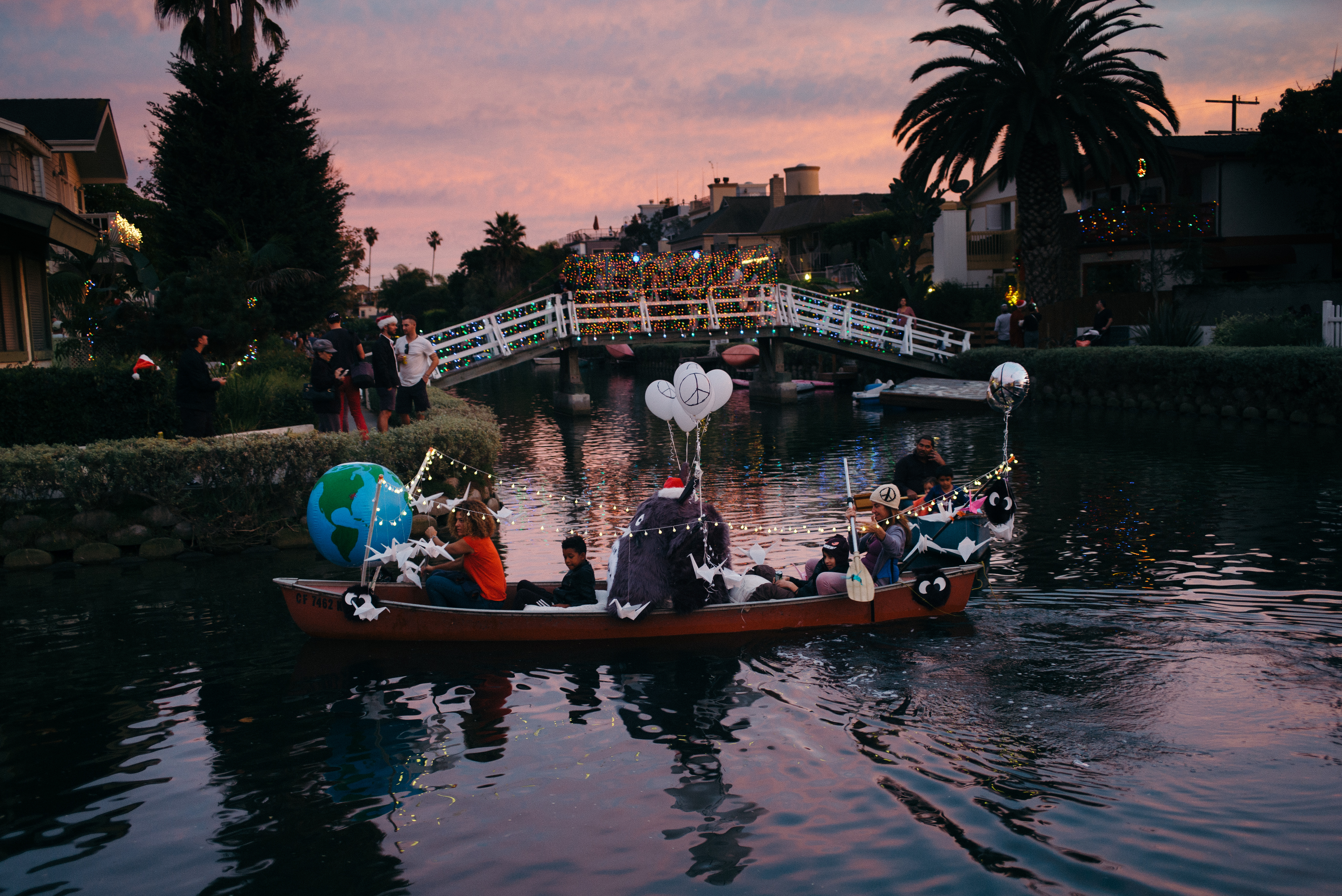 A decorated holiday canoe illuminated with string lights, balloons, and an inflatable globe carrying families down the Venice Canals under a pink sunset sky in Los Angeles, shot with a Leica M240.
