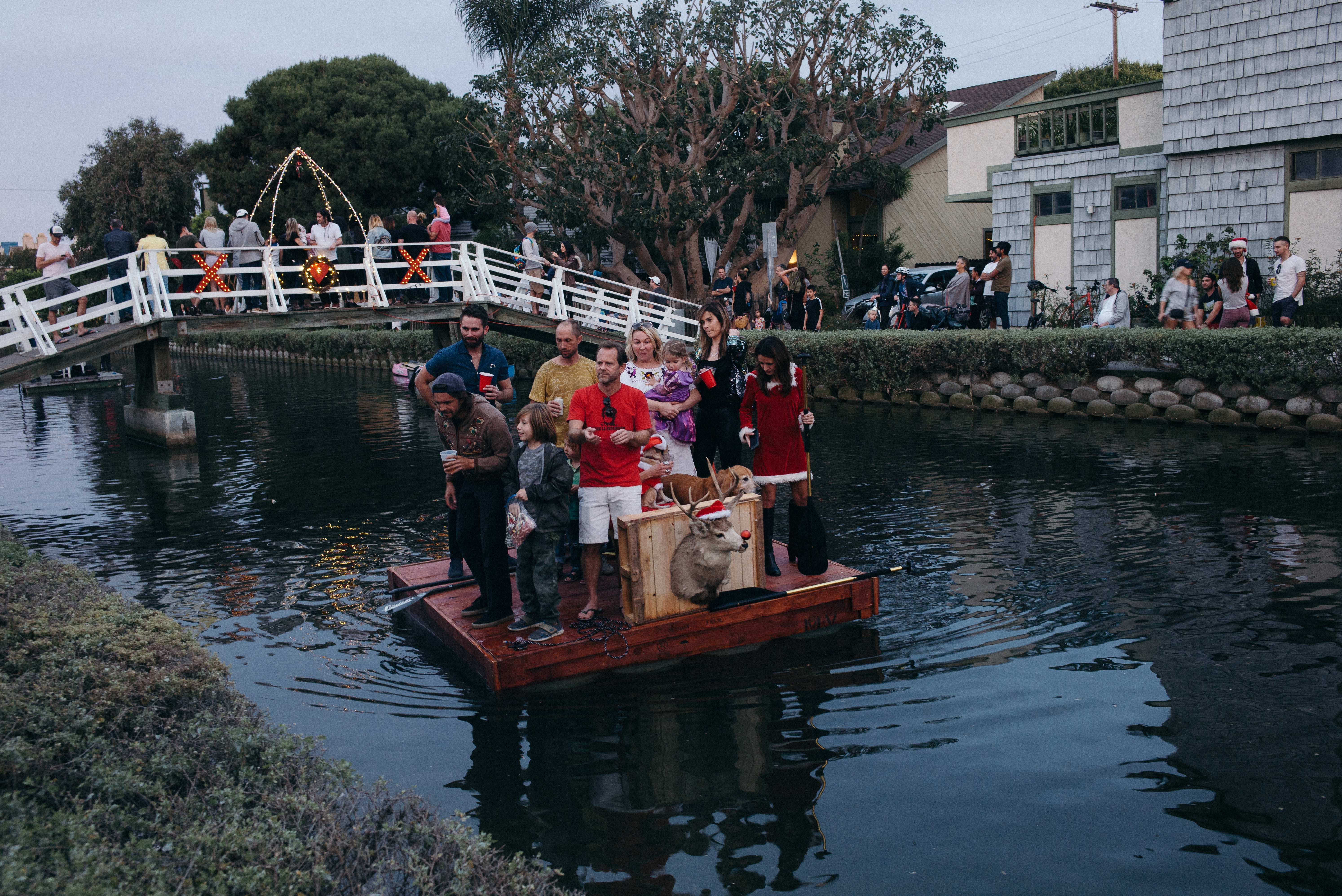 A group of friends, families, and dogs on a wooden holiday raft float passing under a festive bridge crowd during the Venice Canals holiday parade in Los Angeles, shot with a Leica M240.