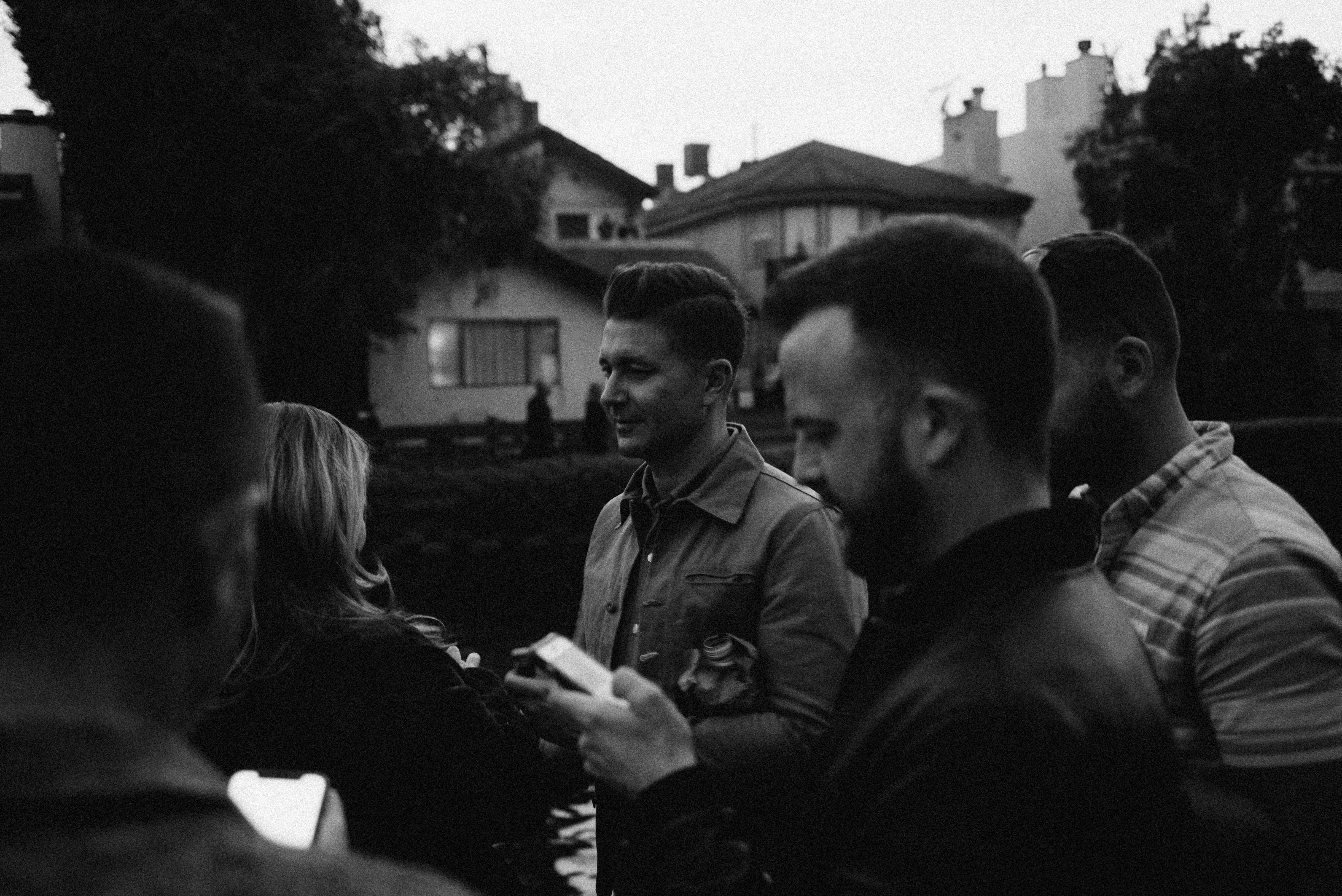 A black and white candid photograph of a group of friends standing outdoors in the evening dusk, focusing on a man in a jacket in conversation with friends holding drinks and a smartphone, shot with a Leica M240.