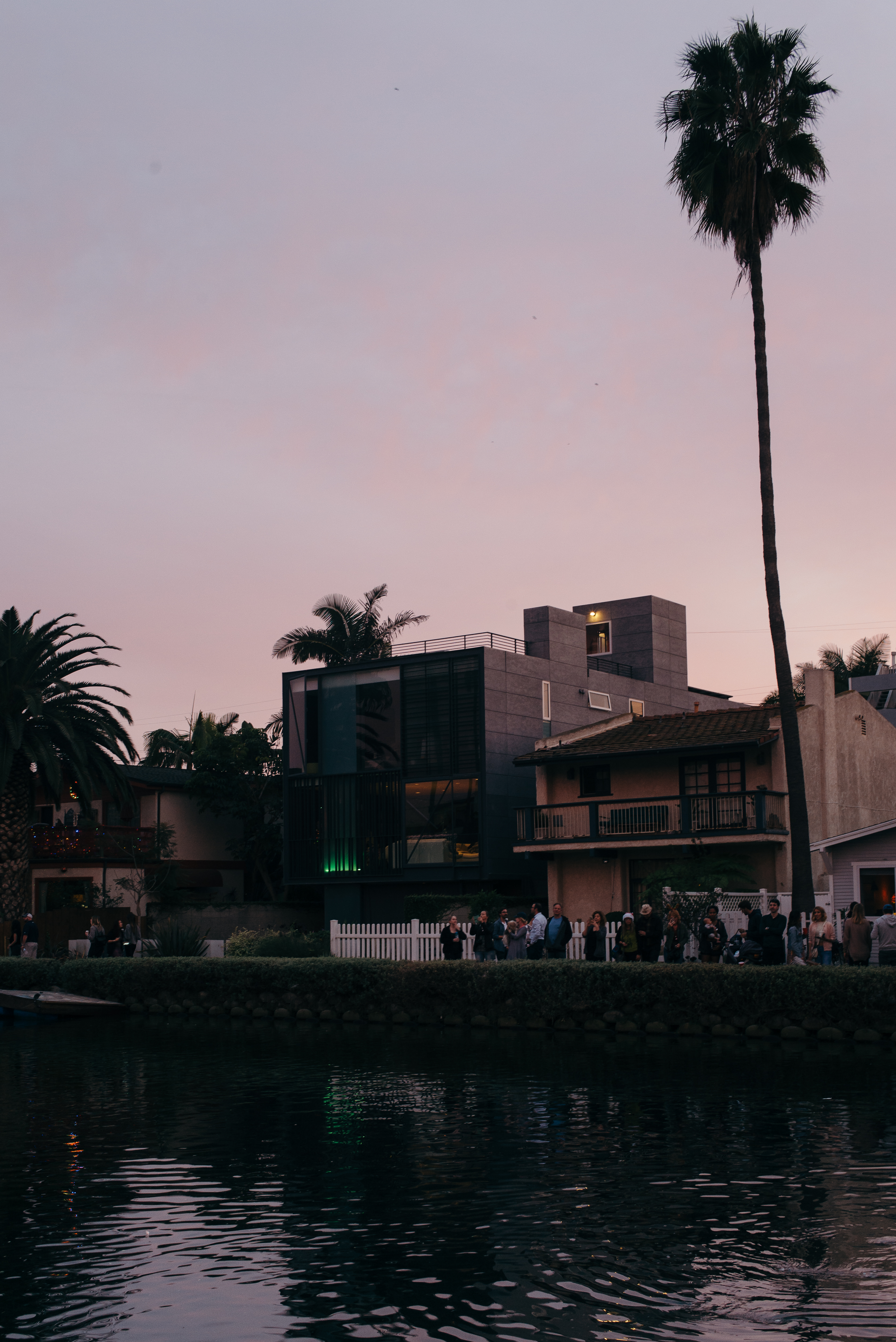 Spectators lining the canal bank and white picket fence in front of modern and rustic canal houses under a soft pink dusk sky with a tall palm tree during the Venice Canals holiday boat parade in Los Angeles, shot with a Leica M240.