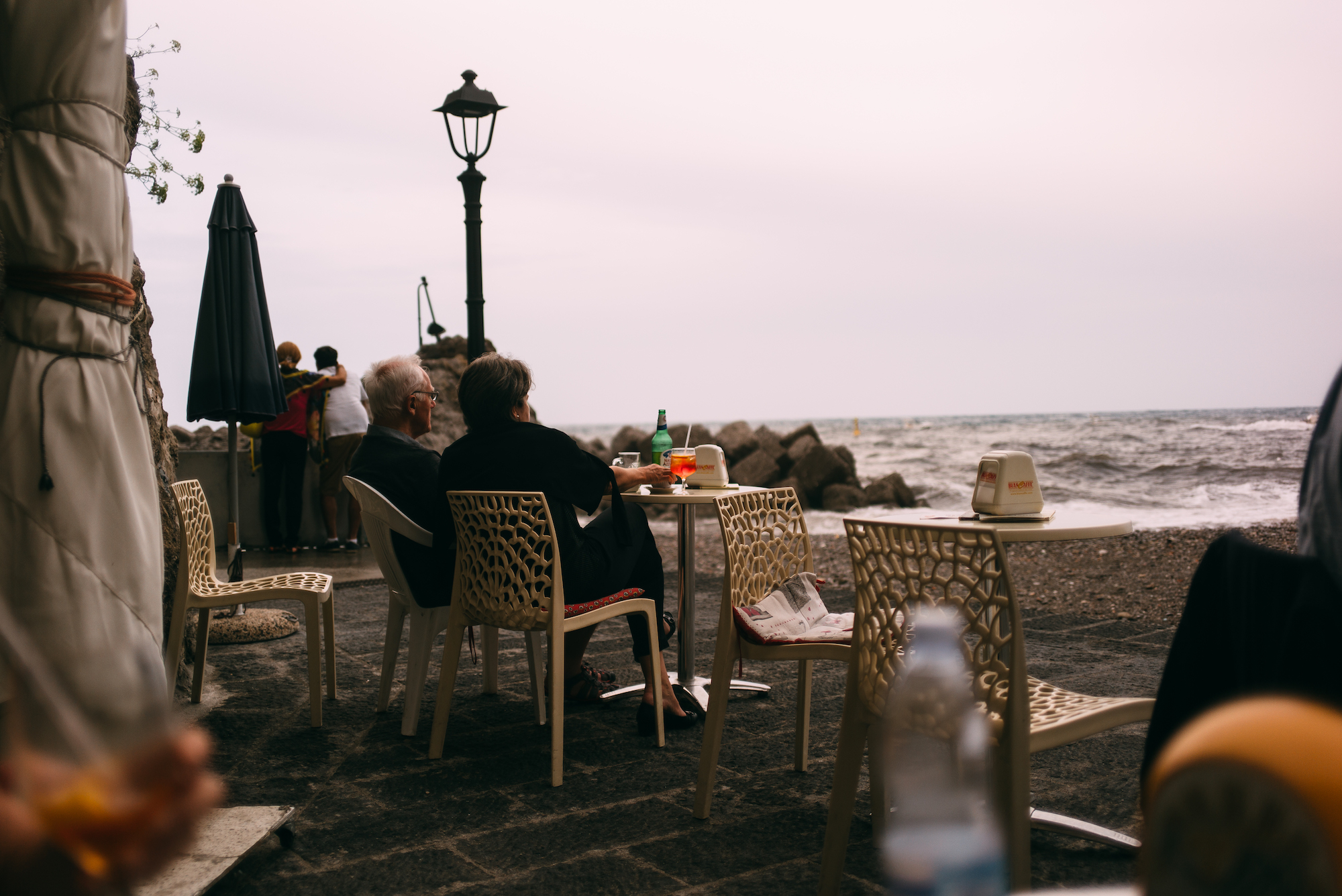 Couple seated at a cafe terrace enjoying drinks before scenic Mediterranean cliffs along the Amalfi Coast.