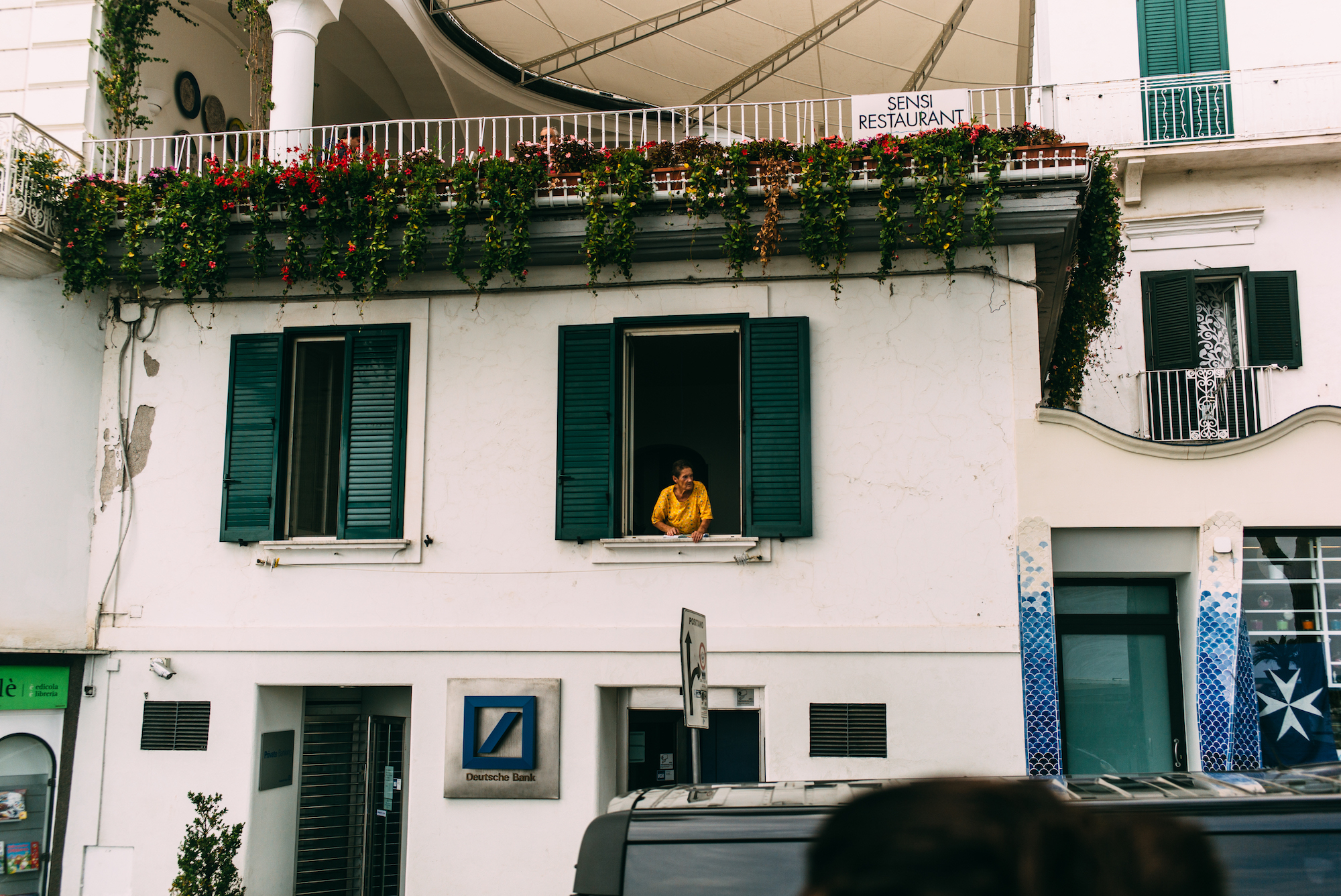Woman leaning casually out of an upper window above a Deutsche Bank branch on an Amalfi Coast street.