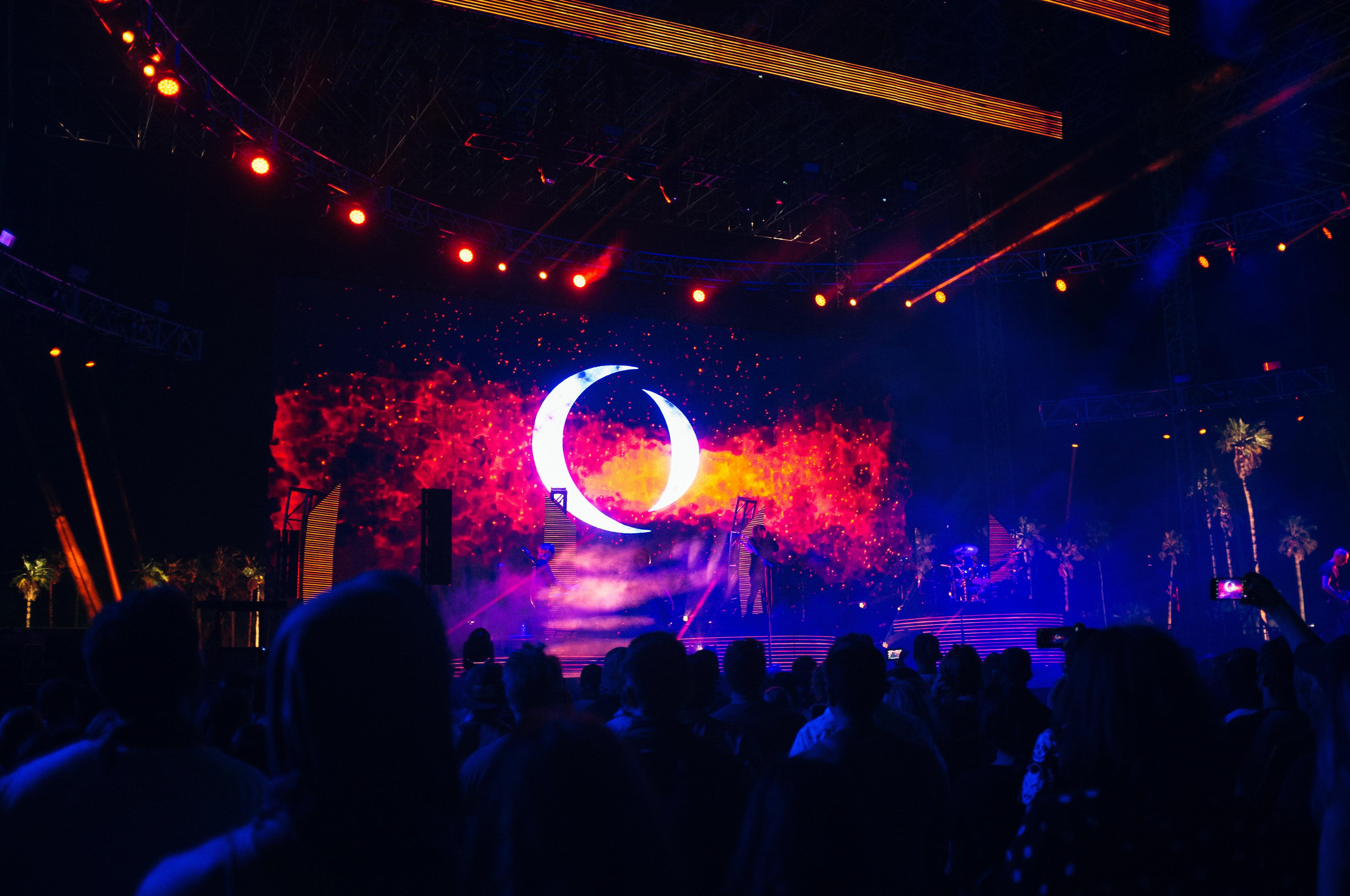Concert stage in deep blue light featuring a giant glowing crescent moon backdrop and musicians at Coachella.