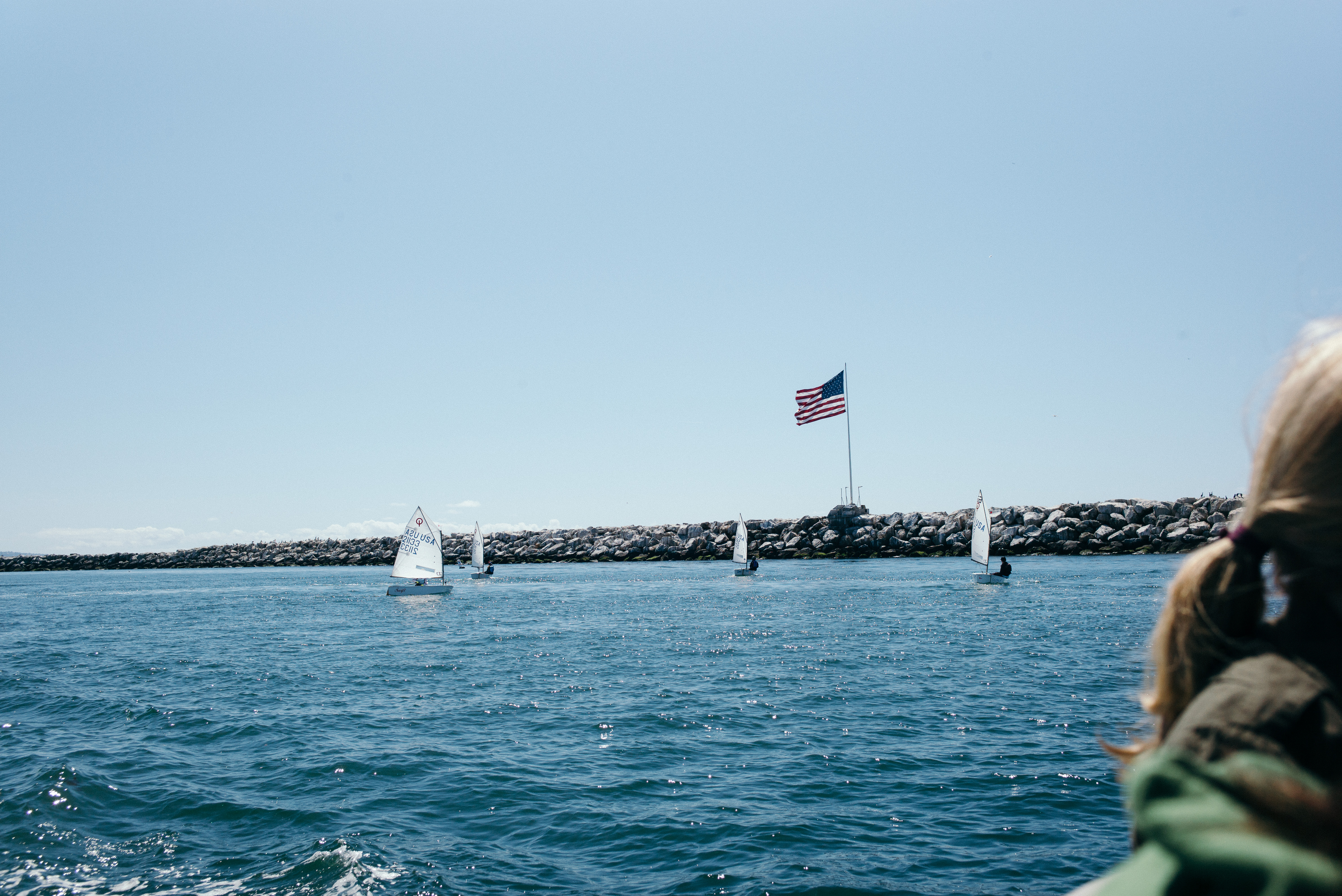 Small sailing dinghies navigating calm harbor waters beside a rocky breakwater flying an American flag.