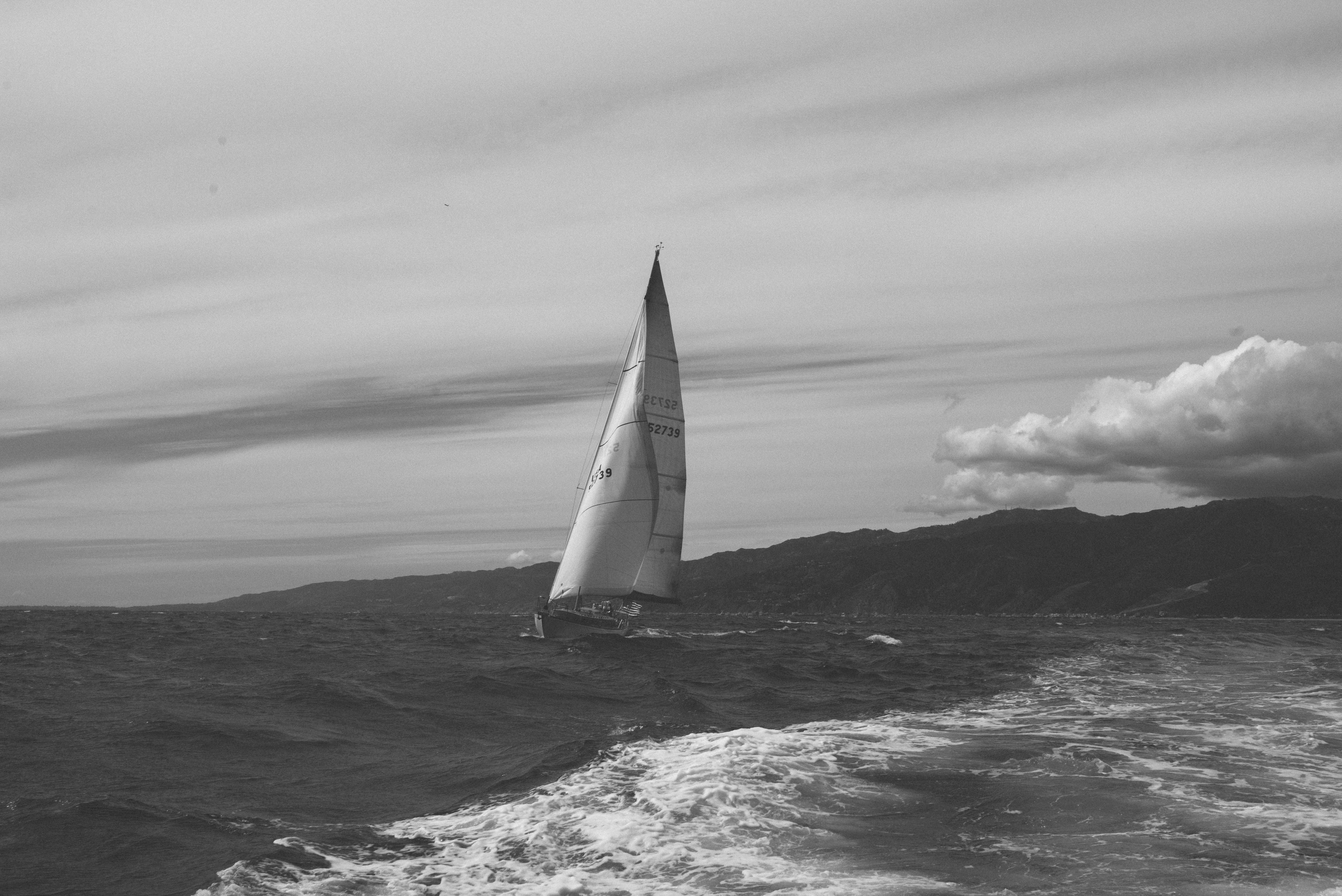 Sailboat heeling under full sail as it cuts through choppy ocean waves against a rugged mountain coast.