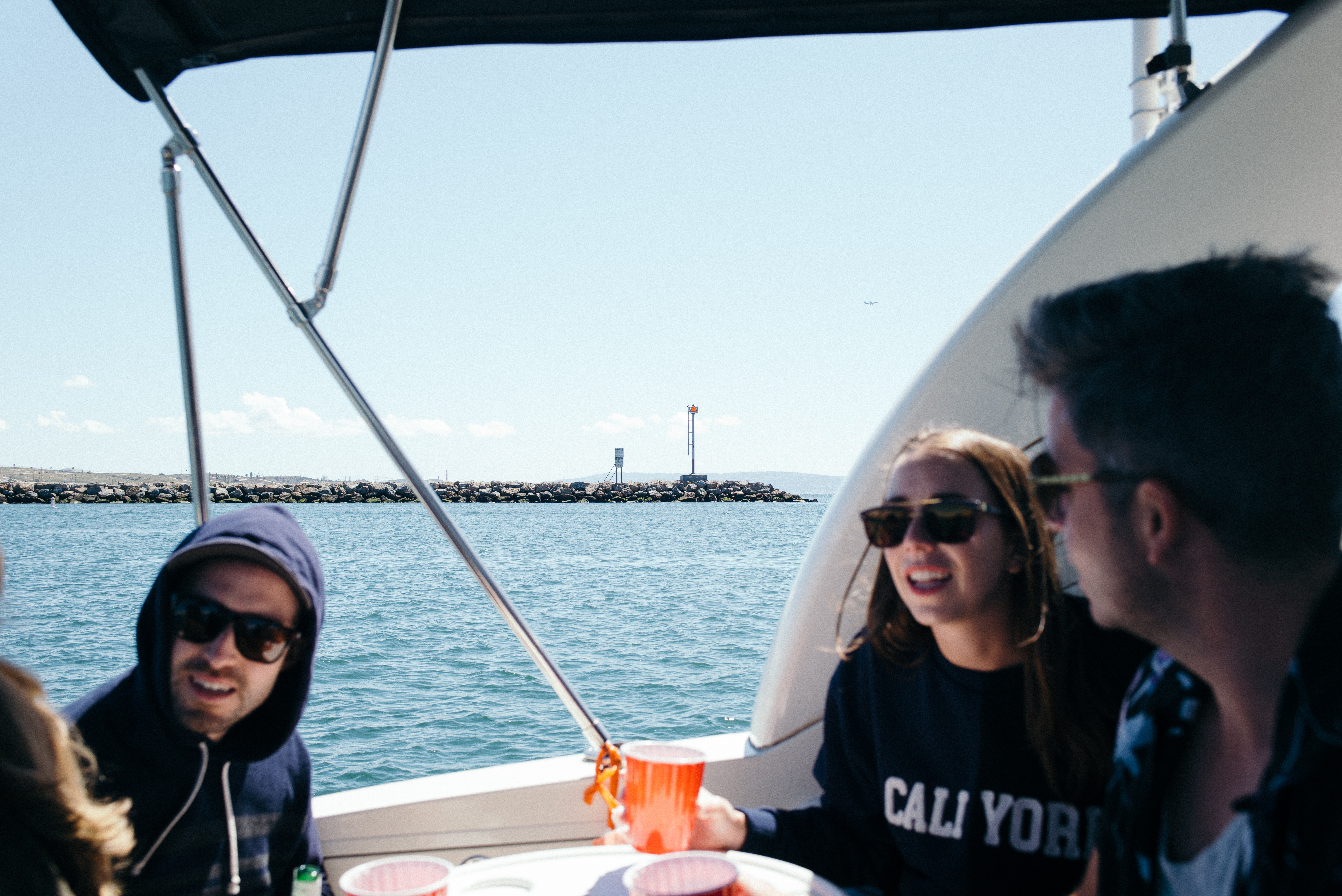 Four smiling friends holding cups in the cockpit of a motorboat on a sunny day near a harbor breakwater.