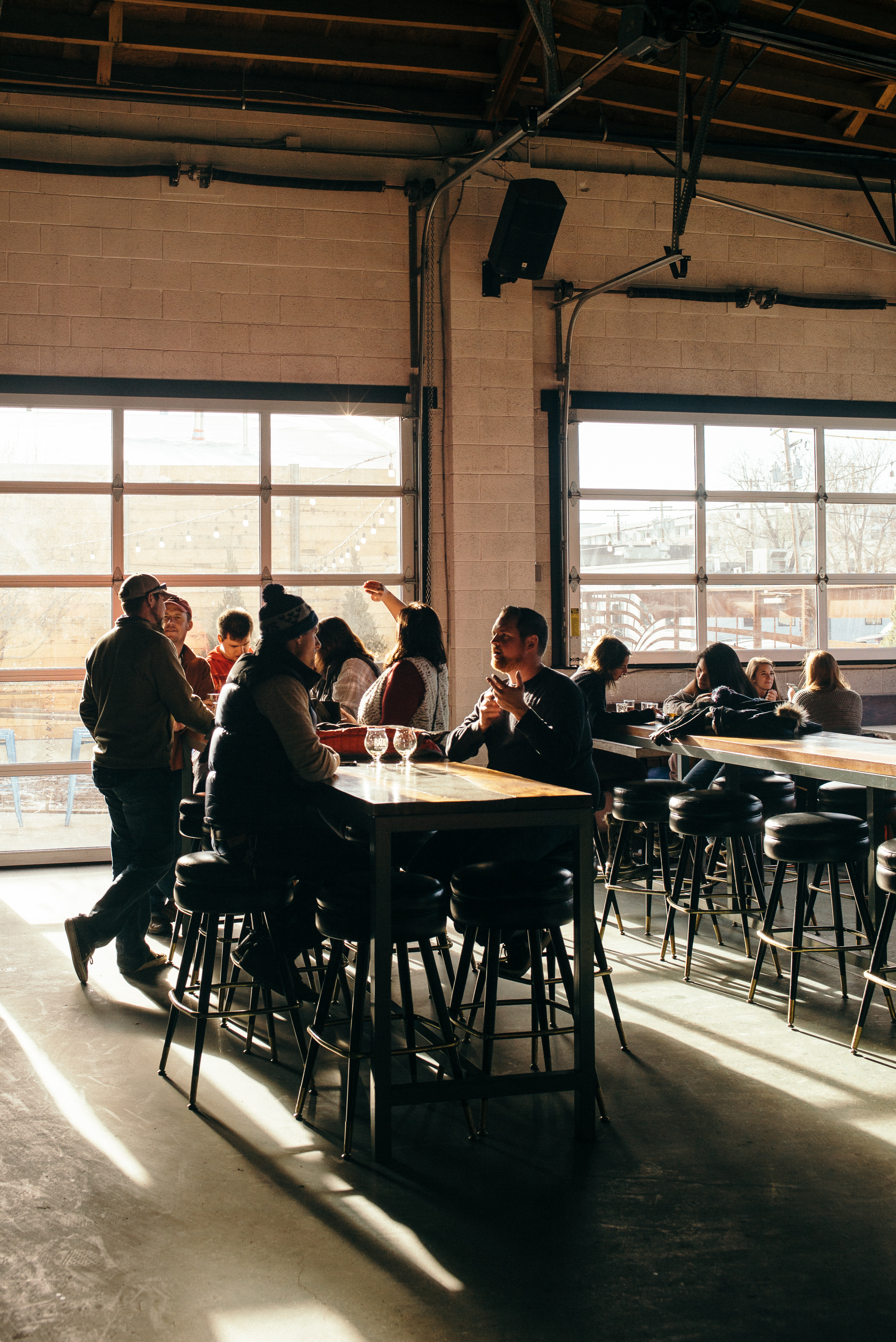 Sunlit brewery taproom with roll-up garage doors, high ceilings, and patrons at a long wooden table.