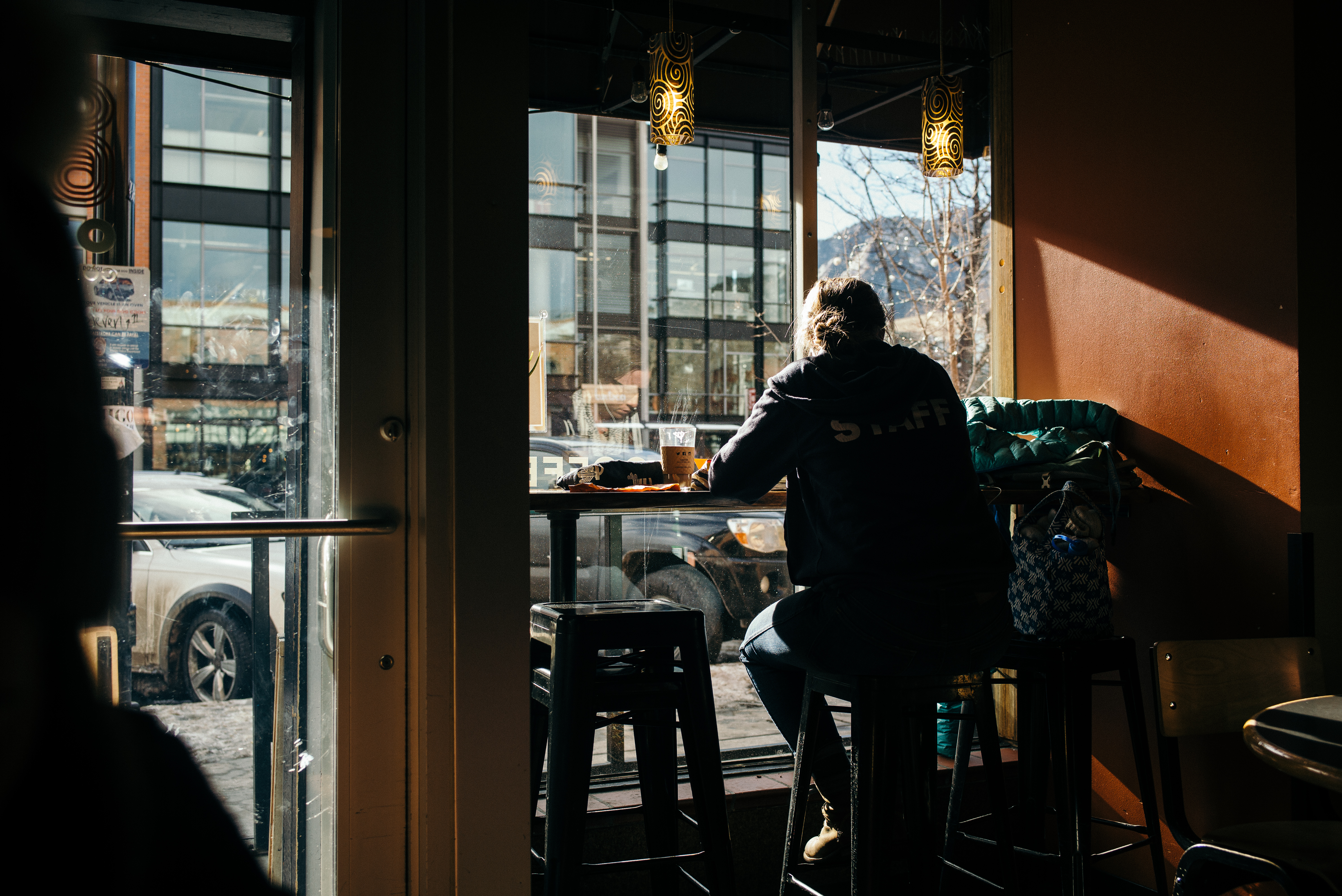 Person in a 'STAFF' hoodie seated at a window counter looking out at city streets and distant mountains.