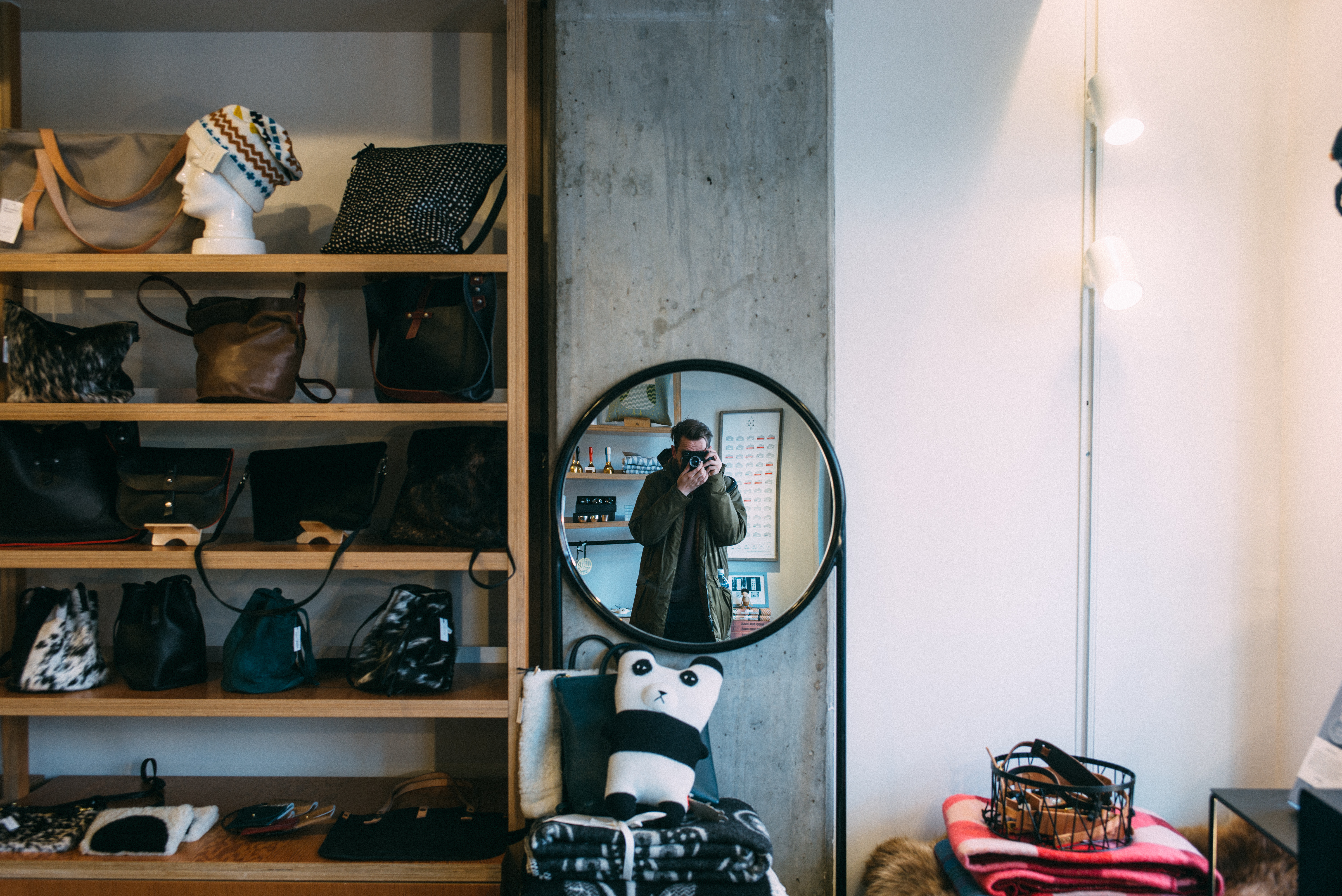 Photographer in an olive parka holding a camera to his face in a mirror inside a leather goods boutique.